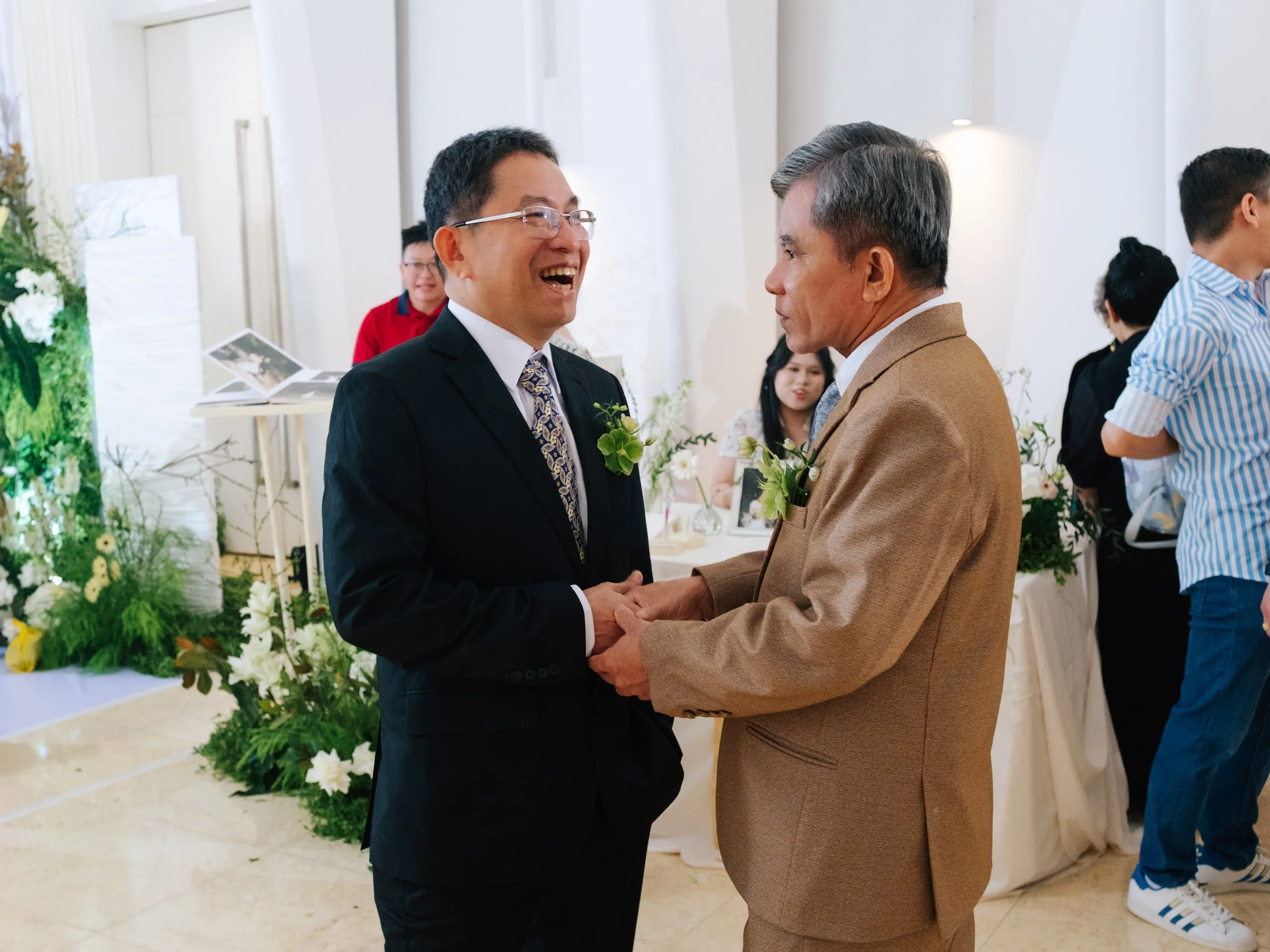 Two men dressed in suits holding hands and smiling at each other in a wedding celebration, surrounded by friends and floral decorations.