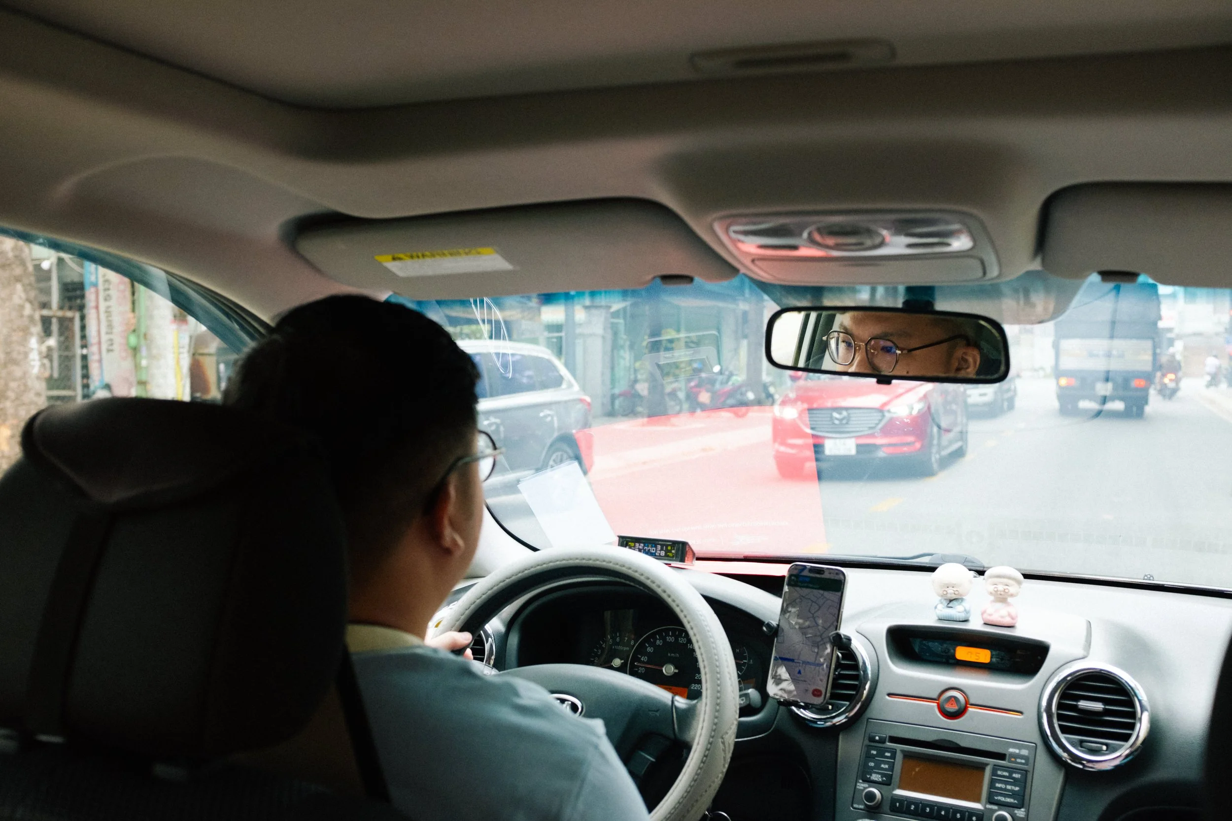 Interior view of a car with a driver wearing glasses, looking at the road. The car's dashboard and rearview mirror are visible, along with two small figurines on the dashboard. The front view through the windshield shows a street with parked and movi