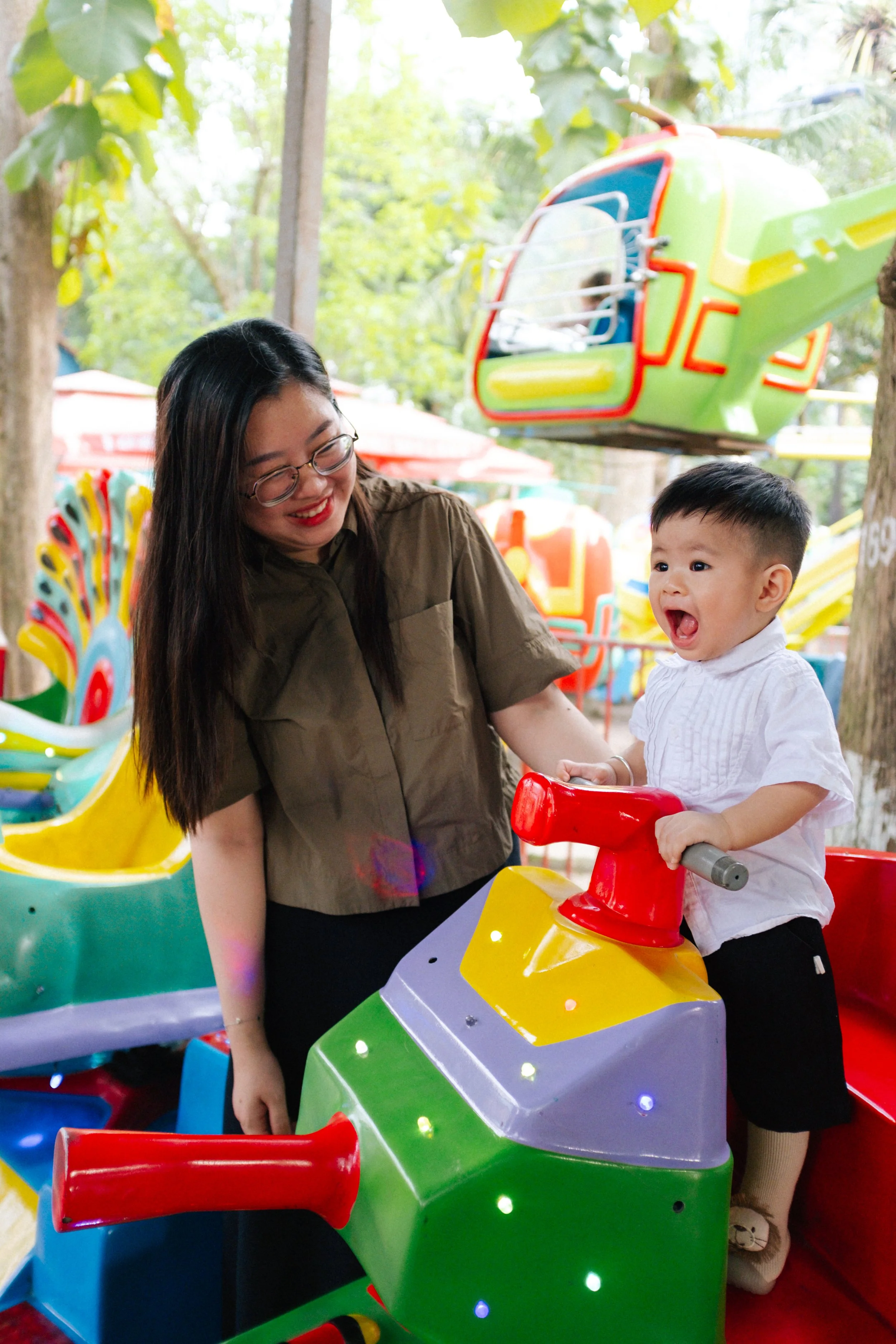 A woman and a young boy enjoying a ride on a colorful amusement park train ride, with trees and amusement rides in the background.