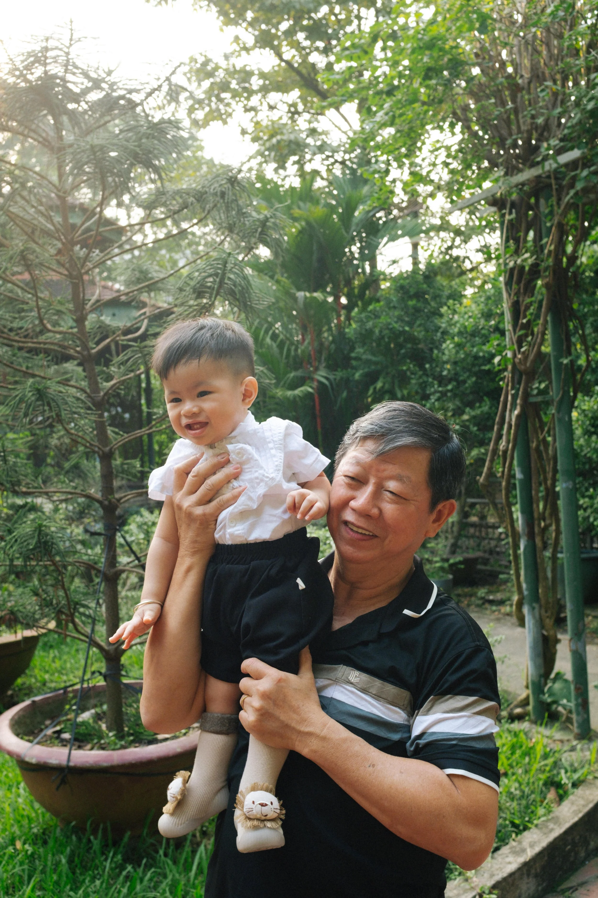 An elderly man holding a young girl in an outdoor garden with trees and plants in the background.