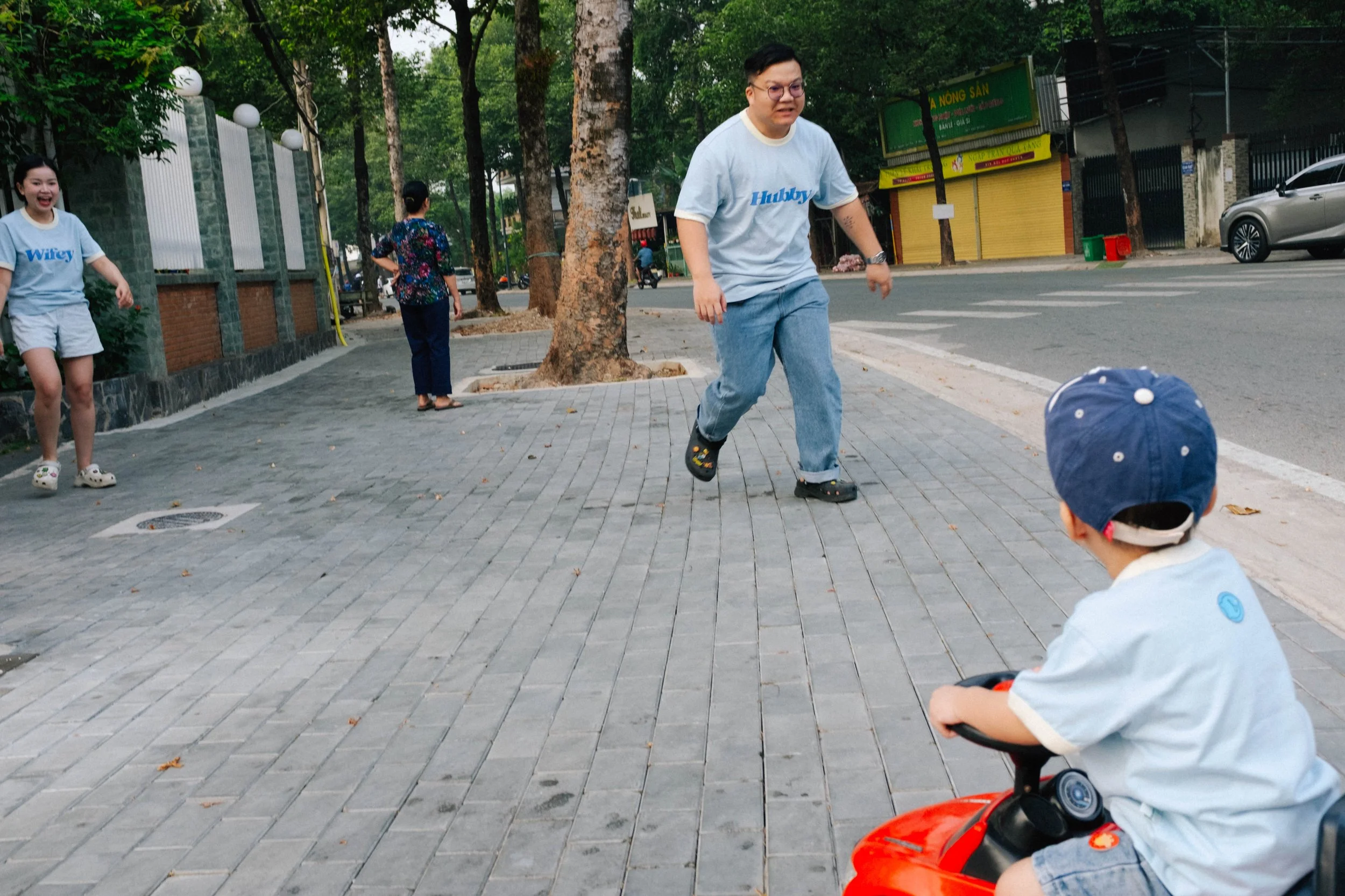 A family playing on the sidewalk, with a man running towards a young boy on a toy car, and a girl and two women nearby, in an urban street lined with trees.