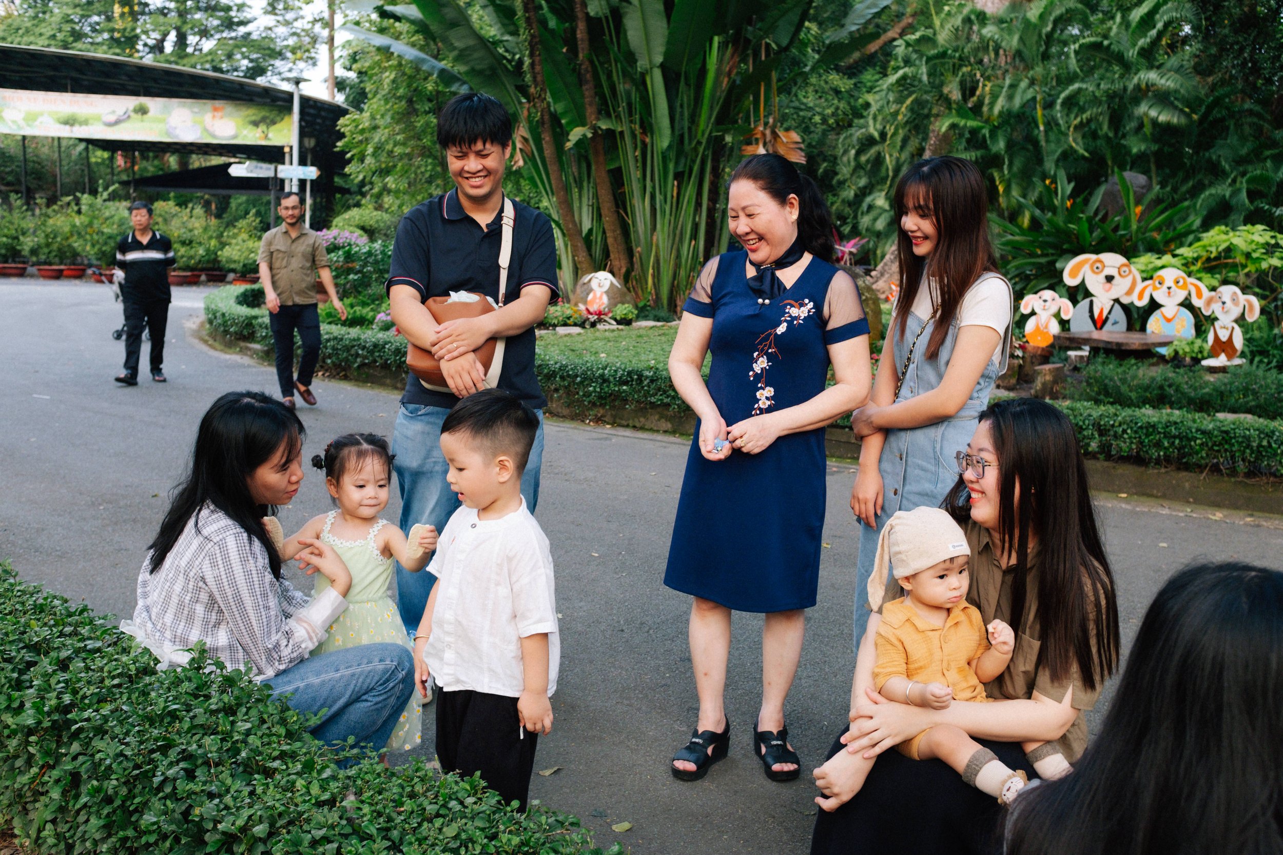 Group of people smiling and interacting outdoors on a paved path, with children sitting and standing, surrounded by lush green plants and cartoon dog decorations in the background.