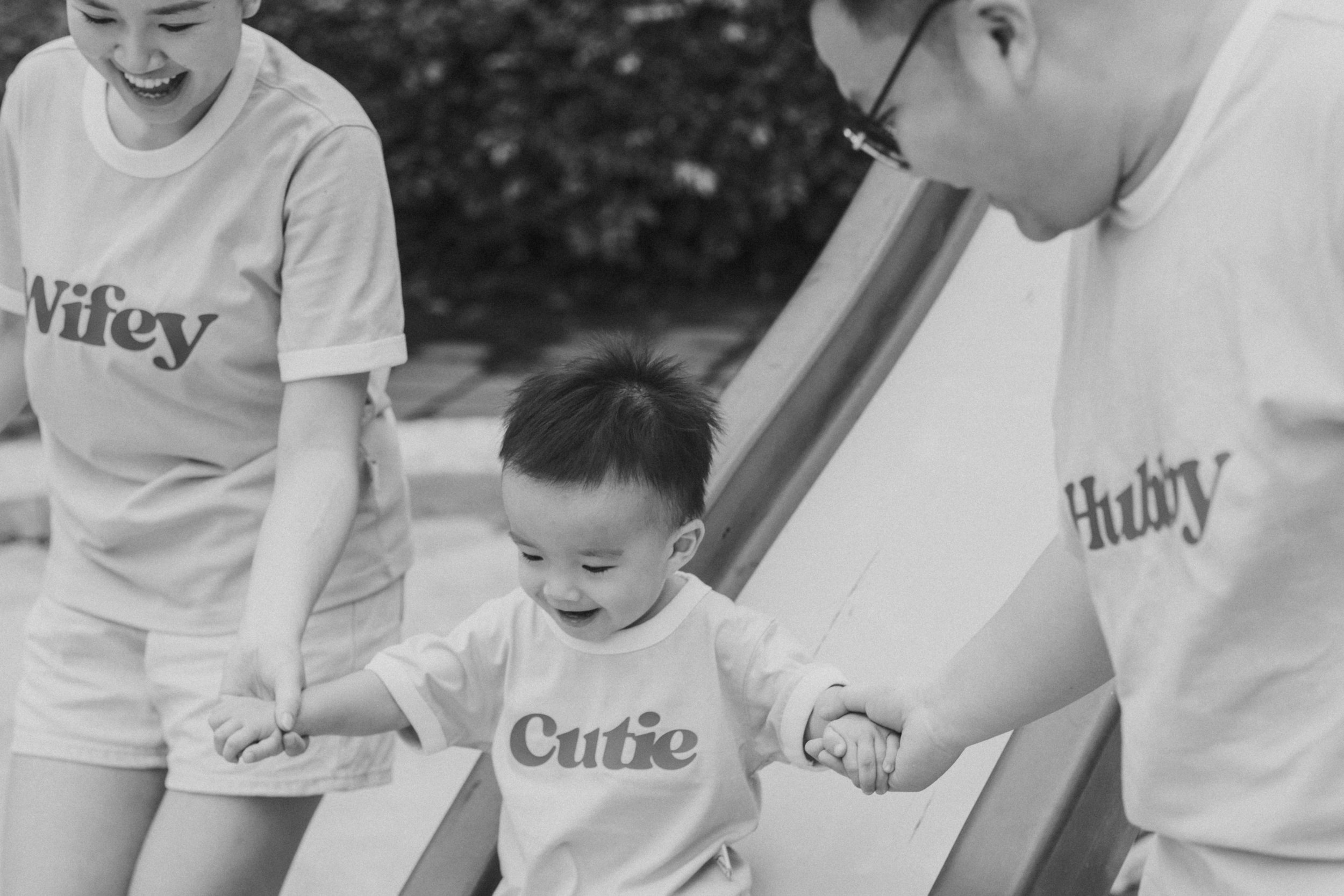 A young child smiling while holding hands with two adults, all wearing matching T-shirts with words 'Cutie' and 'Hubby.'