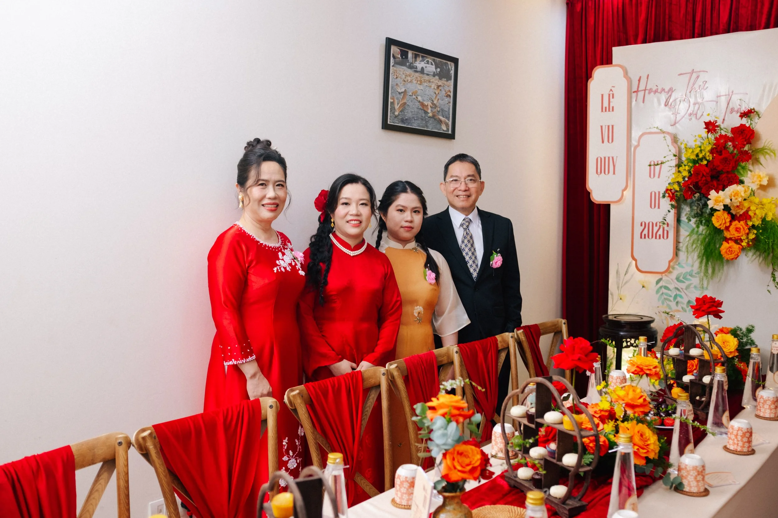 Group of four people dressed in traditional and formal attire standing behind a decorated table at a celebration event, with a backdrop featuring flowers and event details.