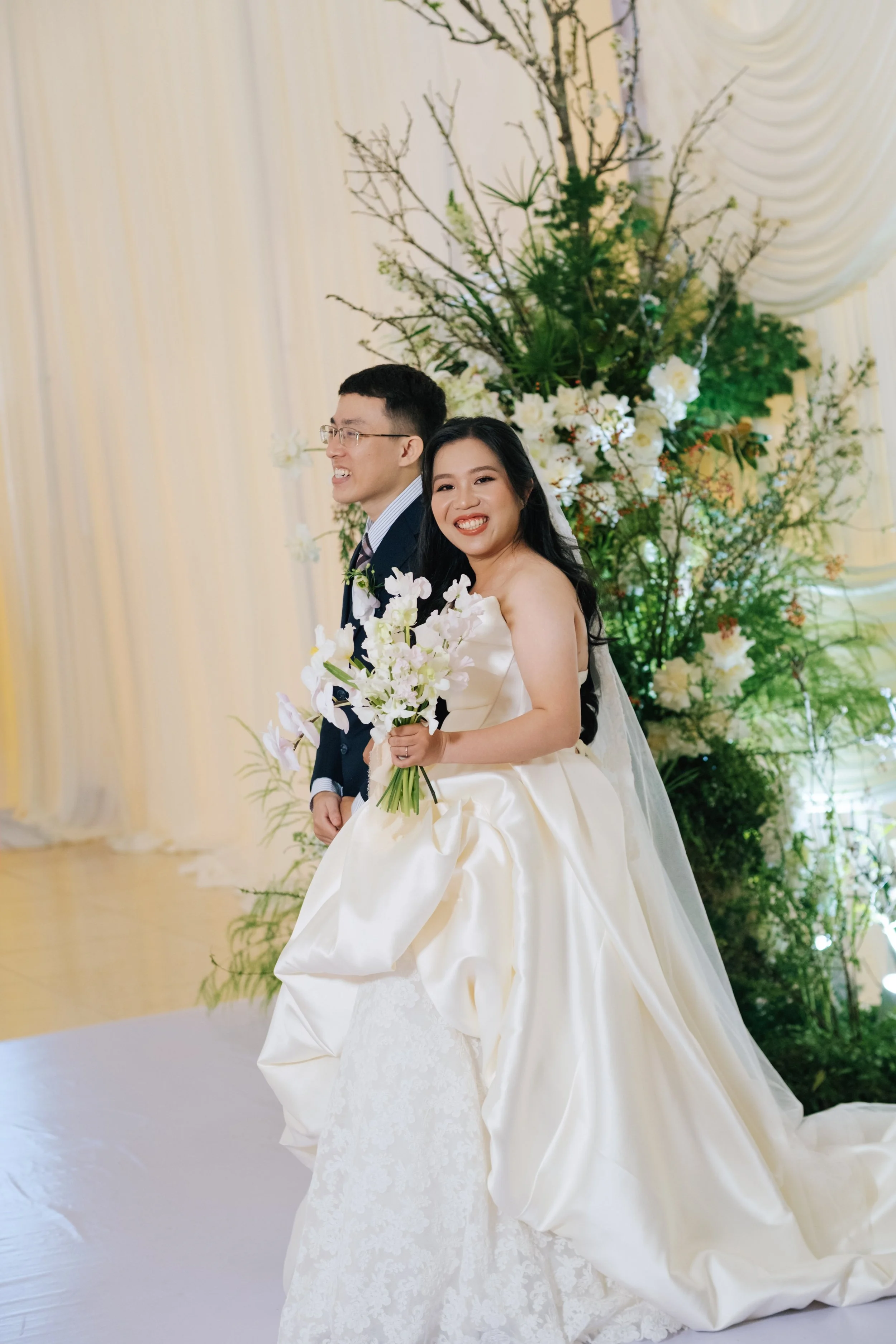 Happy bride in a white wedding gown holding a bouquet standing next to the groom in a suit, with a floral backdrop