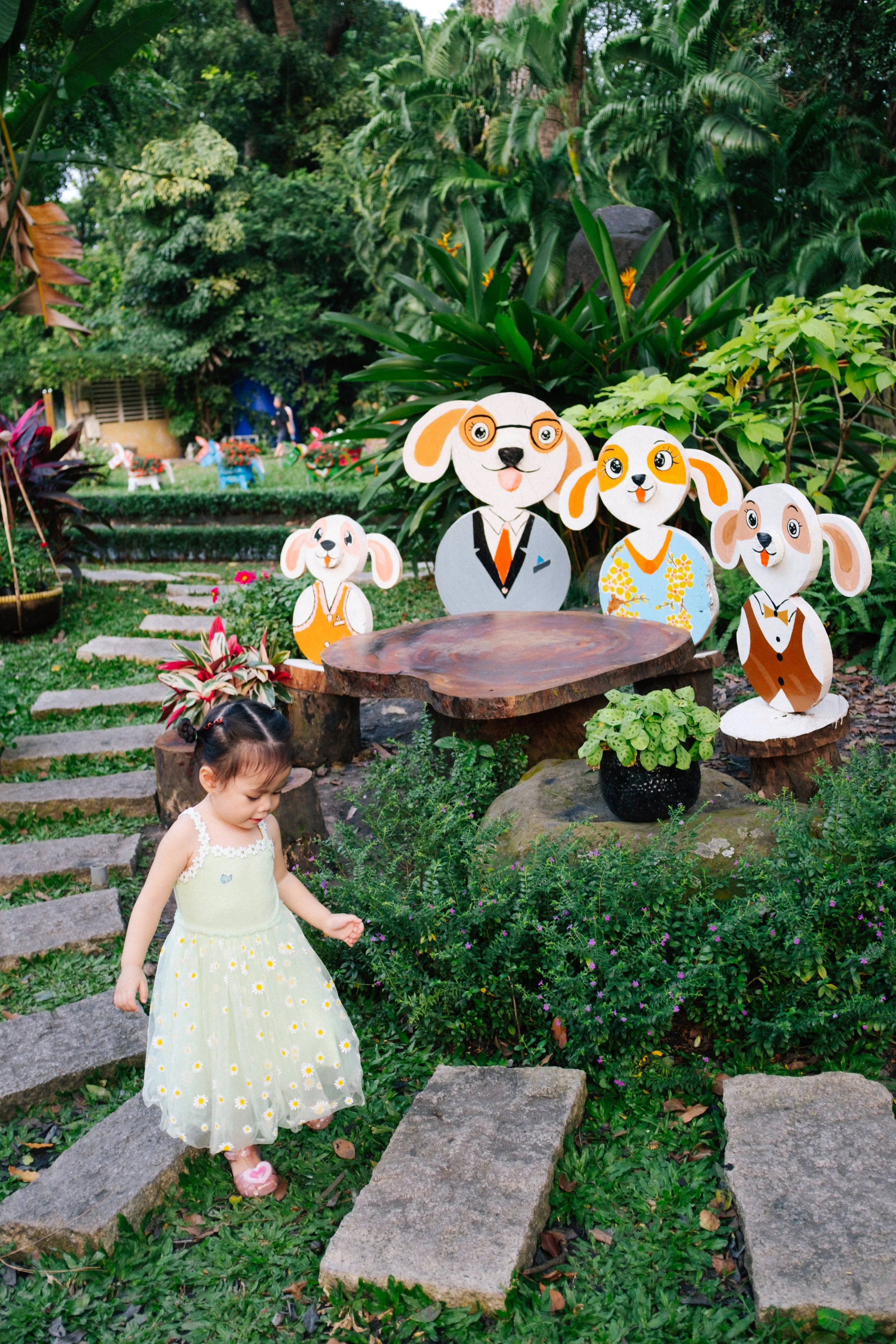 A young girl in a light yellow dress with daisy patterns walking on a stone pathway in a garden with large green plants and trees. Besides her, a real girl with short hair and a flower hairband. Decorative wooden dog cutouts painted with cute dog fac
