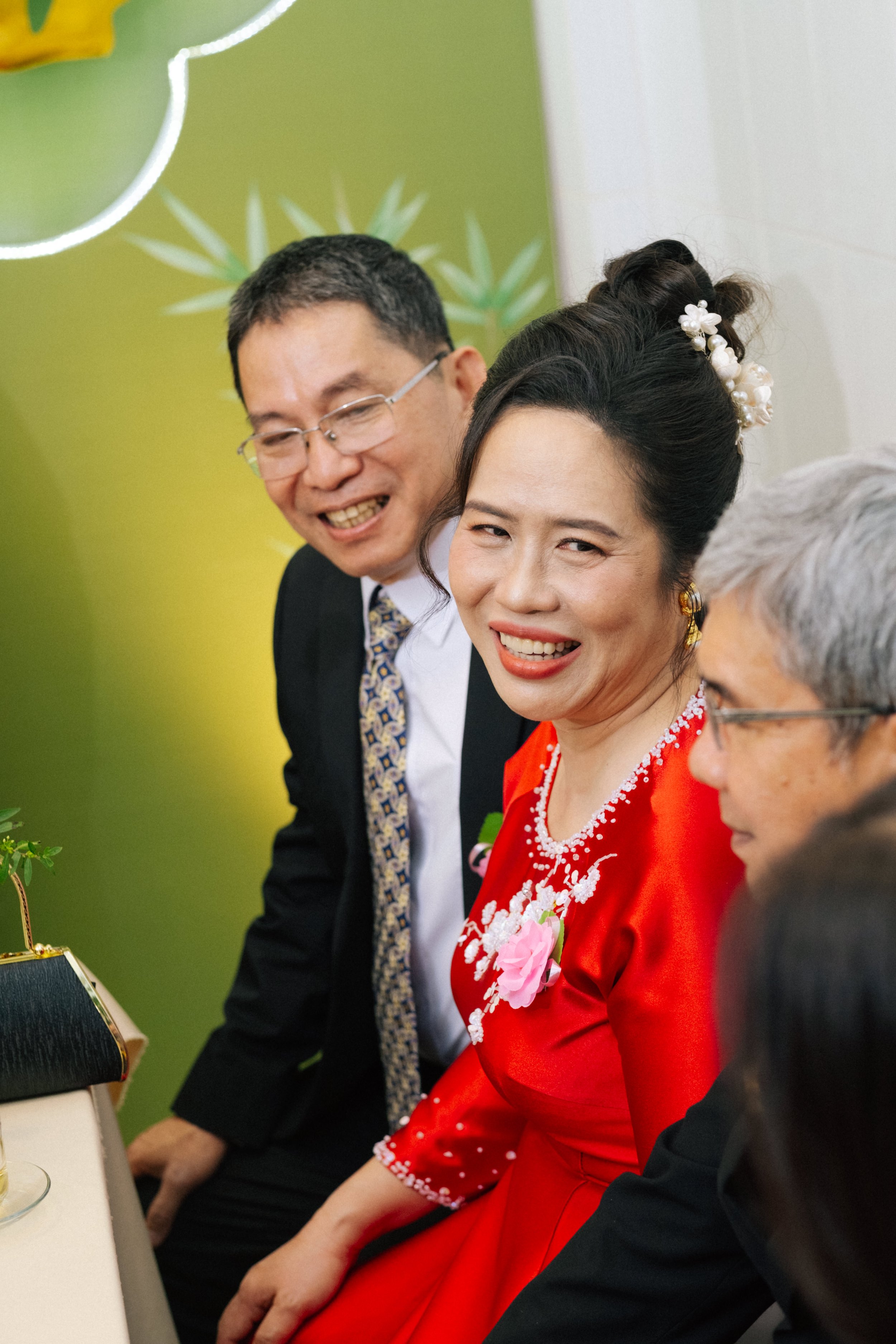 A woman in a red traditional dress with floral embroidery and a woman with a white flower hairpiece, sitting at a table with three other people, all smiling and laughing, at a celebration or gathering