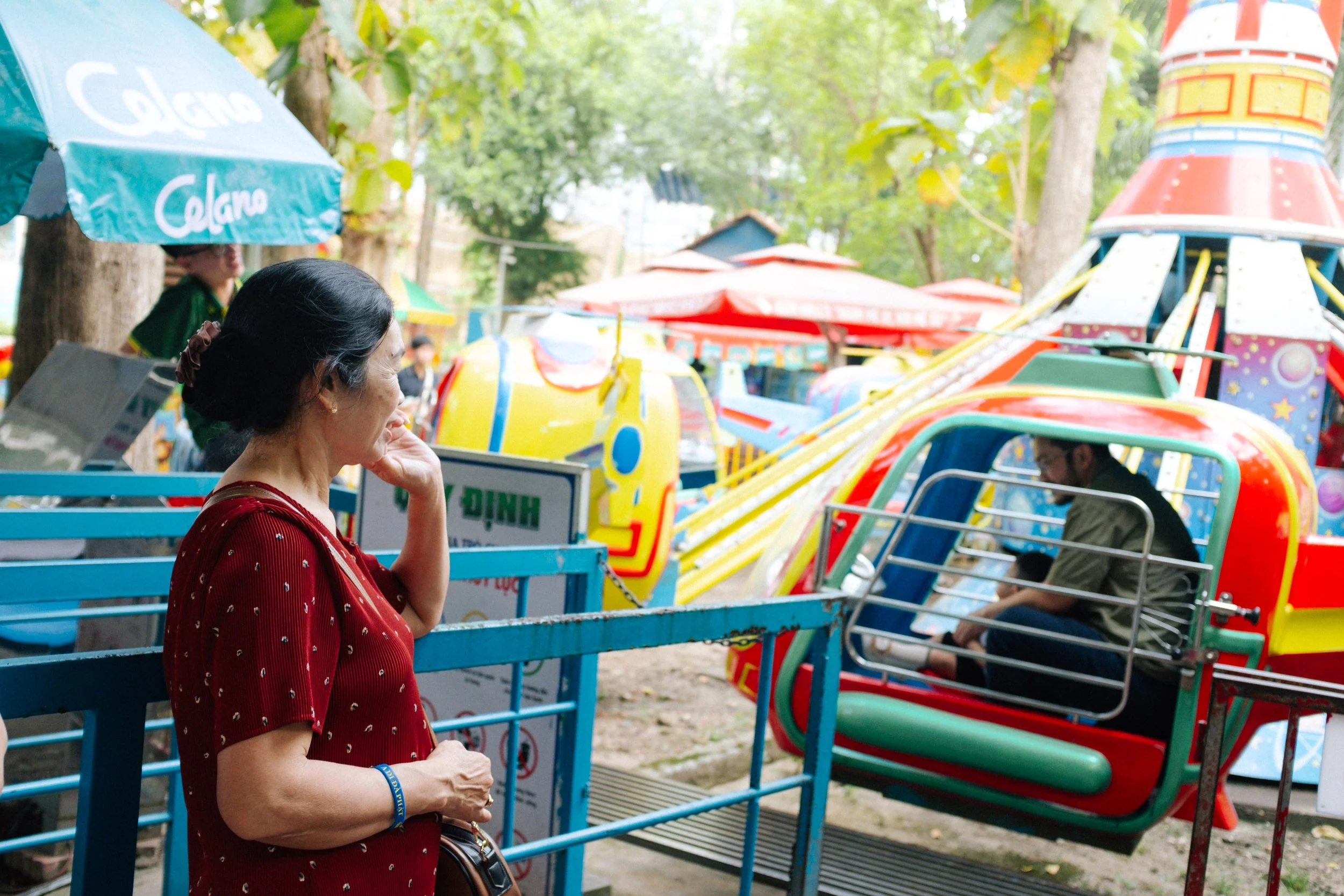 A woman in a red blouse with small white polka dots stands near a carnival ride, looking at a man sitting in a small helicopter ride car surrounded by colorful amusement park attractions and trees in the background.