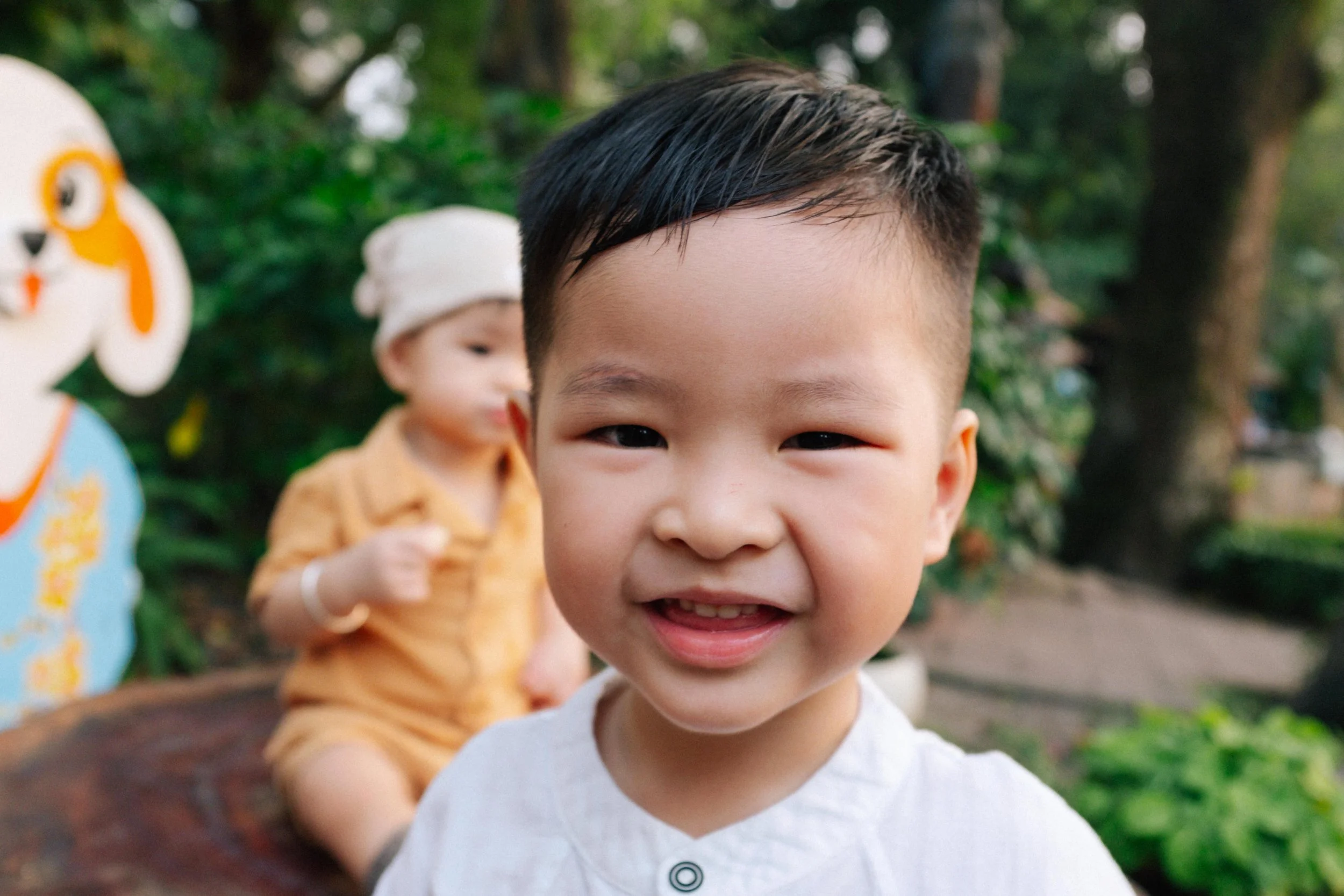 Close-up of a young Asian boy smiling outdoors with a blurred young girl in the background, and colorful cartoon animal decorations on a fence.