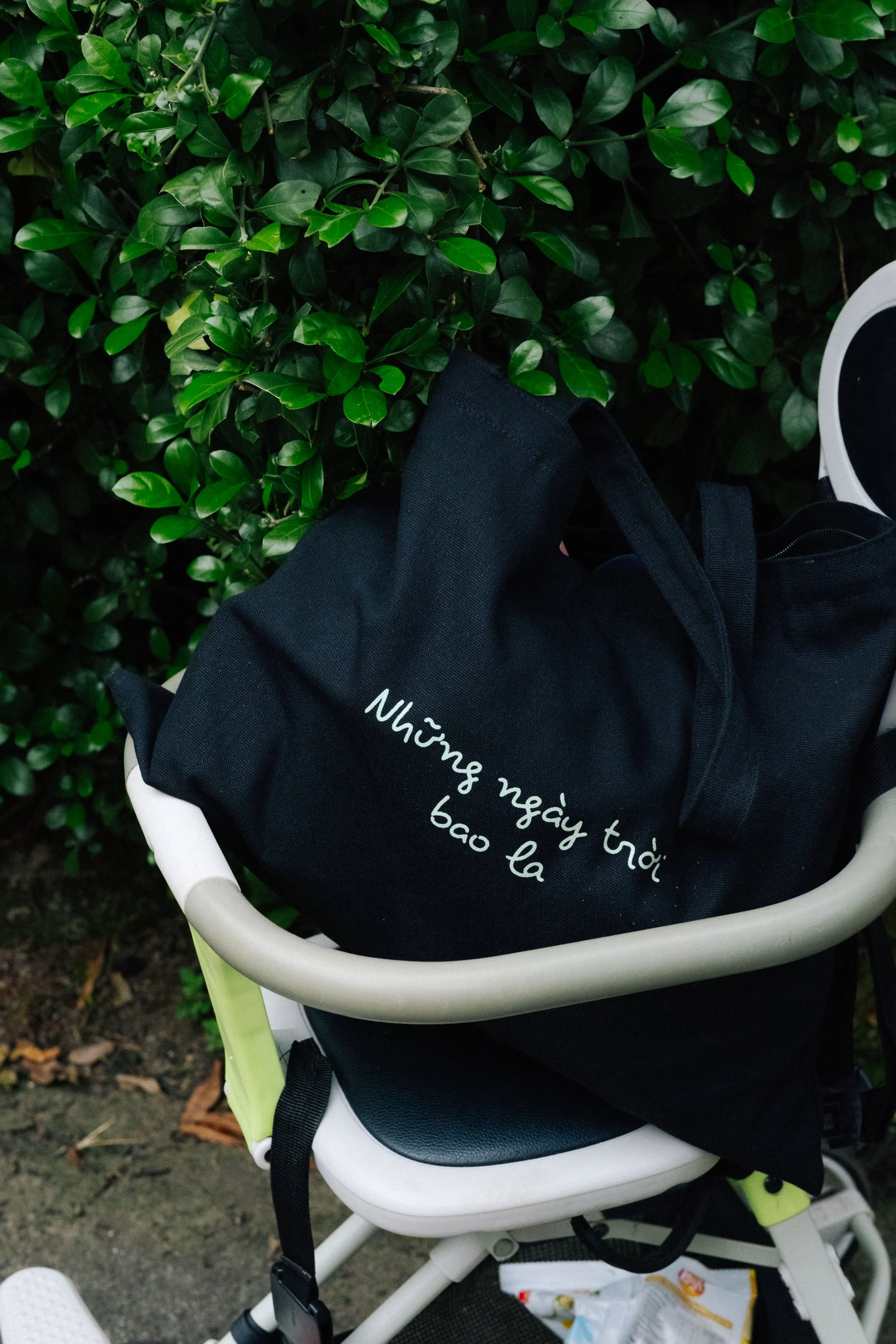 A black bag with white Vietnamese text resting in a gray and white stroller, with green leaves in the background.