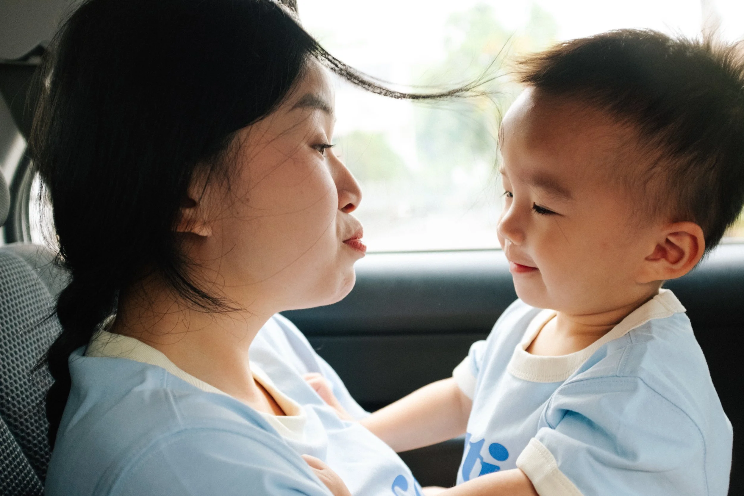A woman and a young boy sit closely in a car, facing each other, with their faces very close, smiling and looking at each other affectionately.