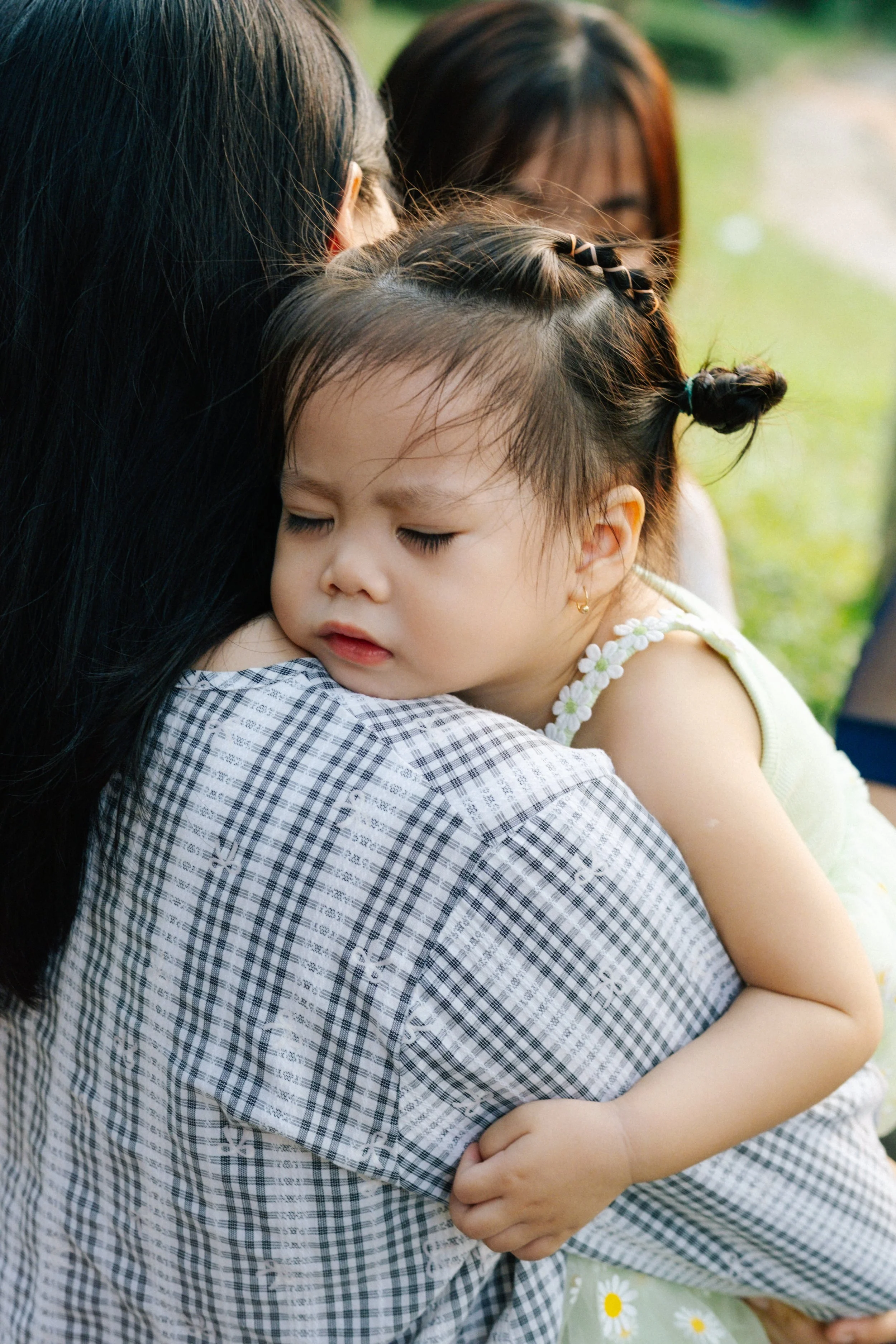 A young girl is sleeping on an adult's shoulder while being hugged, with her eyes closed, outdoors on a grass field.