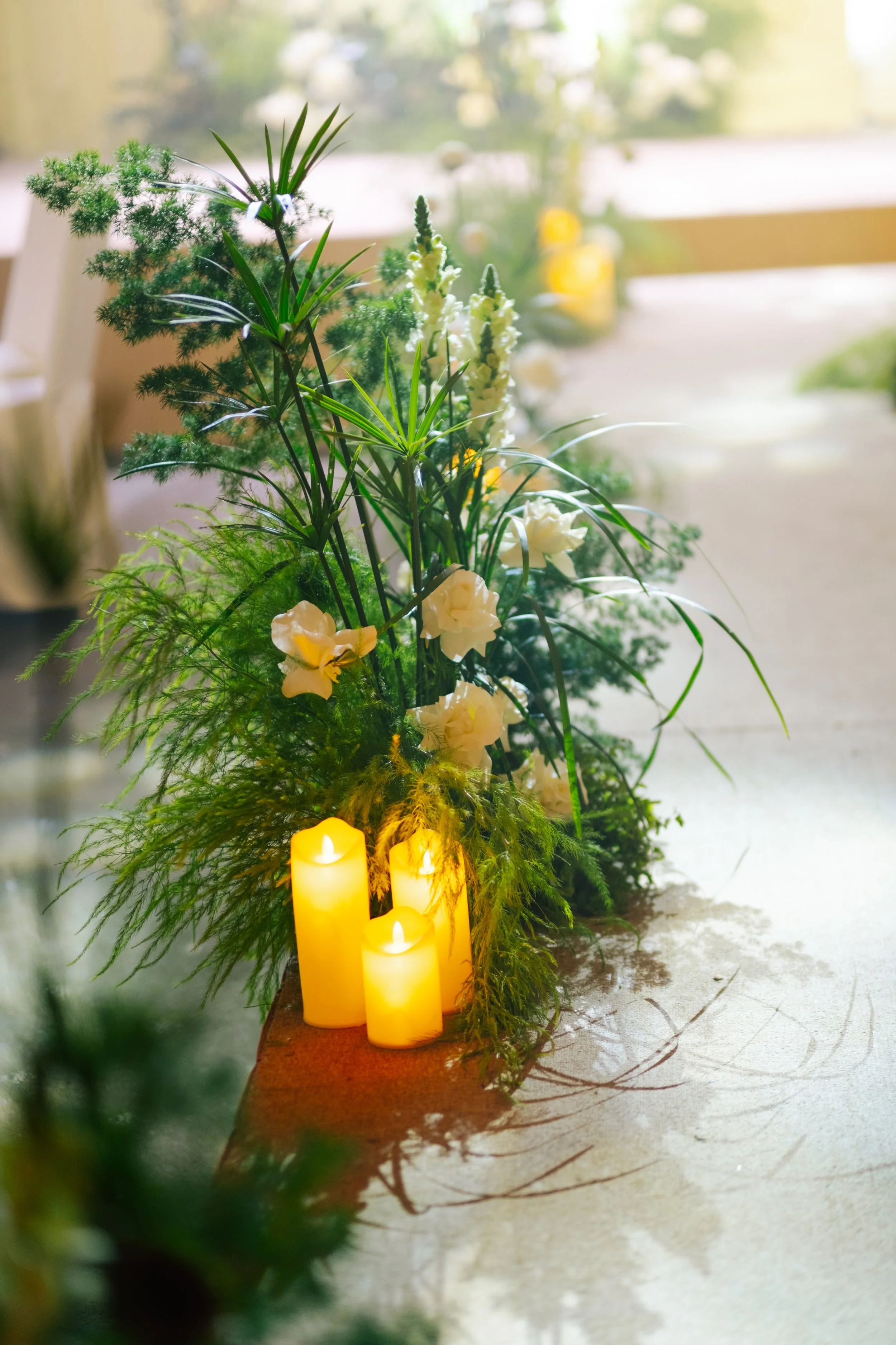 A floral arrangement with white flowers and green foliage, illuminated by three yellow candles, set on a light-colored surface near a window.