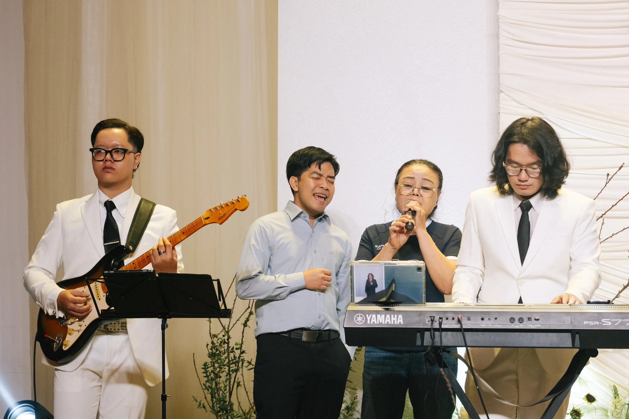 Four people performing music on stage: a guitarist in white suit, a man singing, a woman singing into microphone, and a woman playing keyboard in white suit.