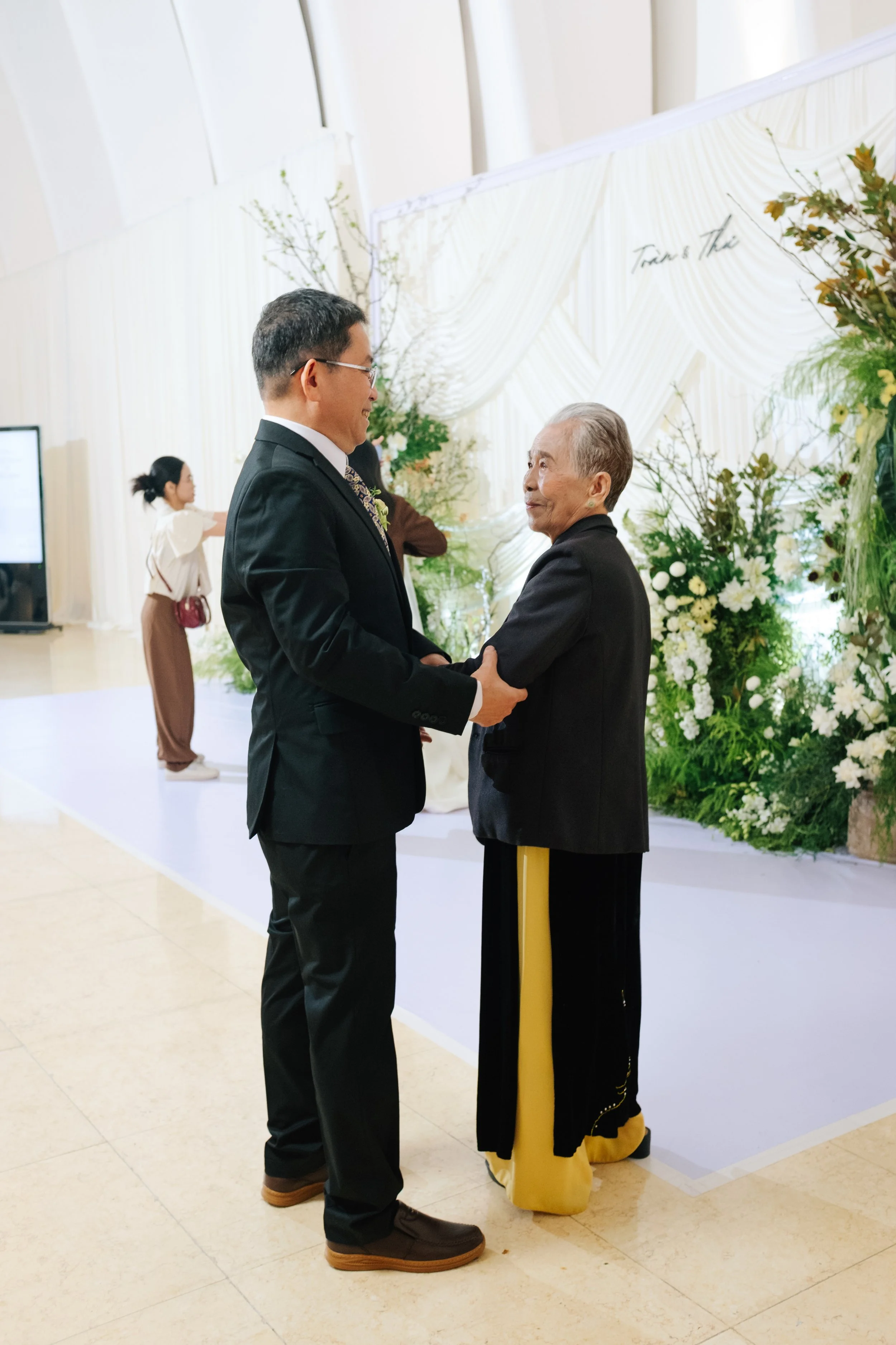 A man and an elderly woman share a moment at a formal event, possibly a wedding, with floral decorations and draped fabric in the background.