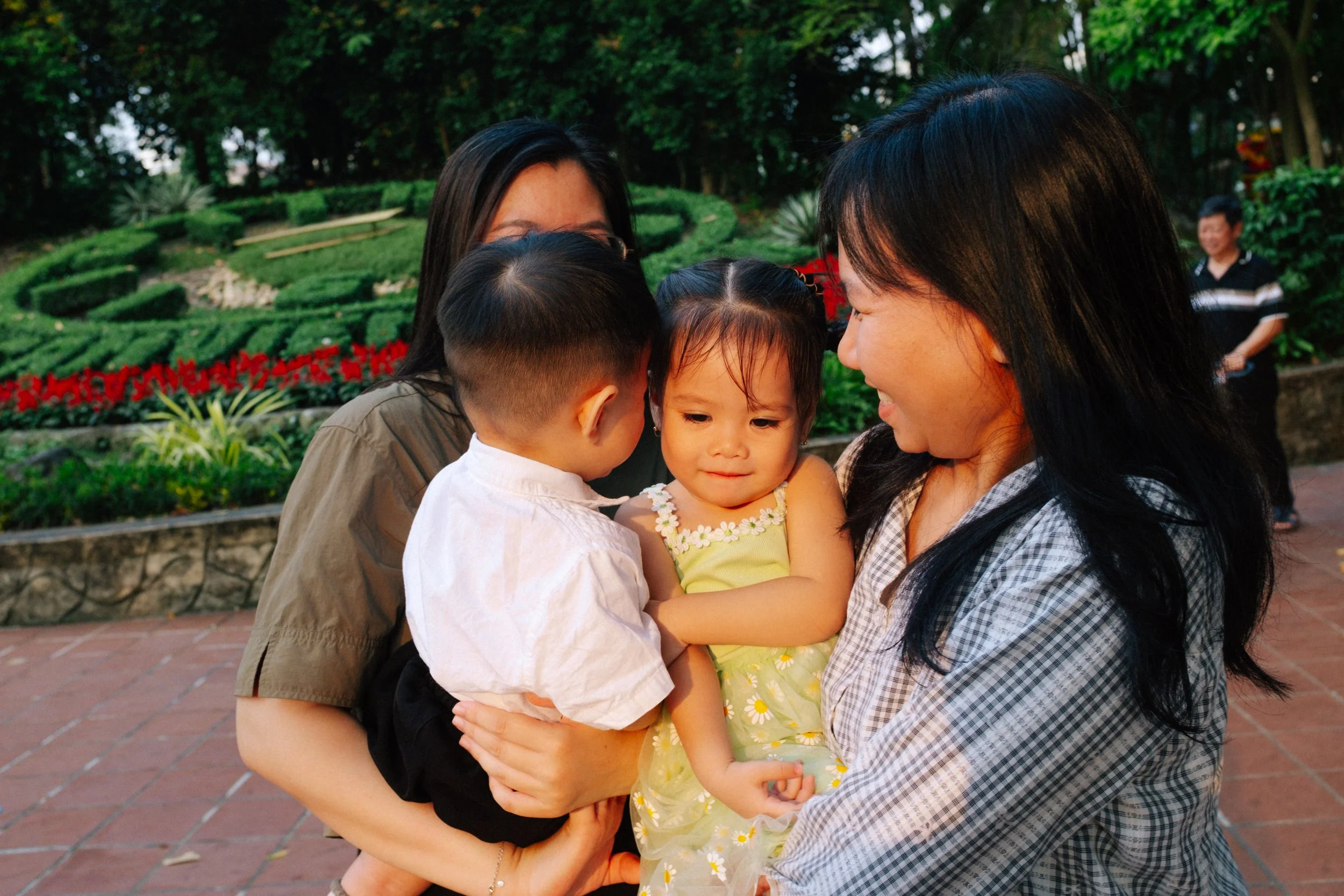 Two women holding two young children, outdoors in a park with greenery and flowers in the background.