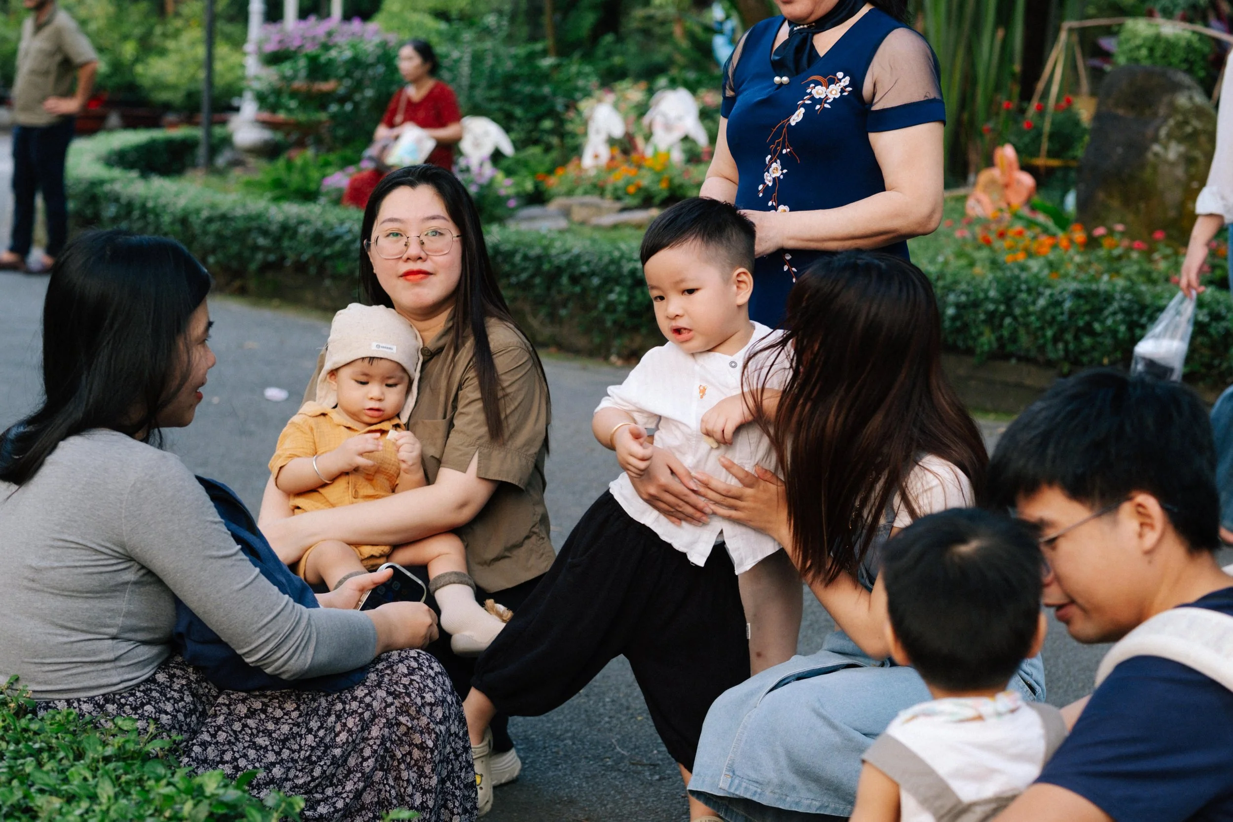 A group of Asian people, including children, gathered outdoors in a garden, engaging in conversation.
