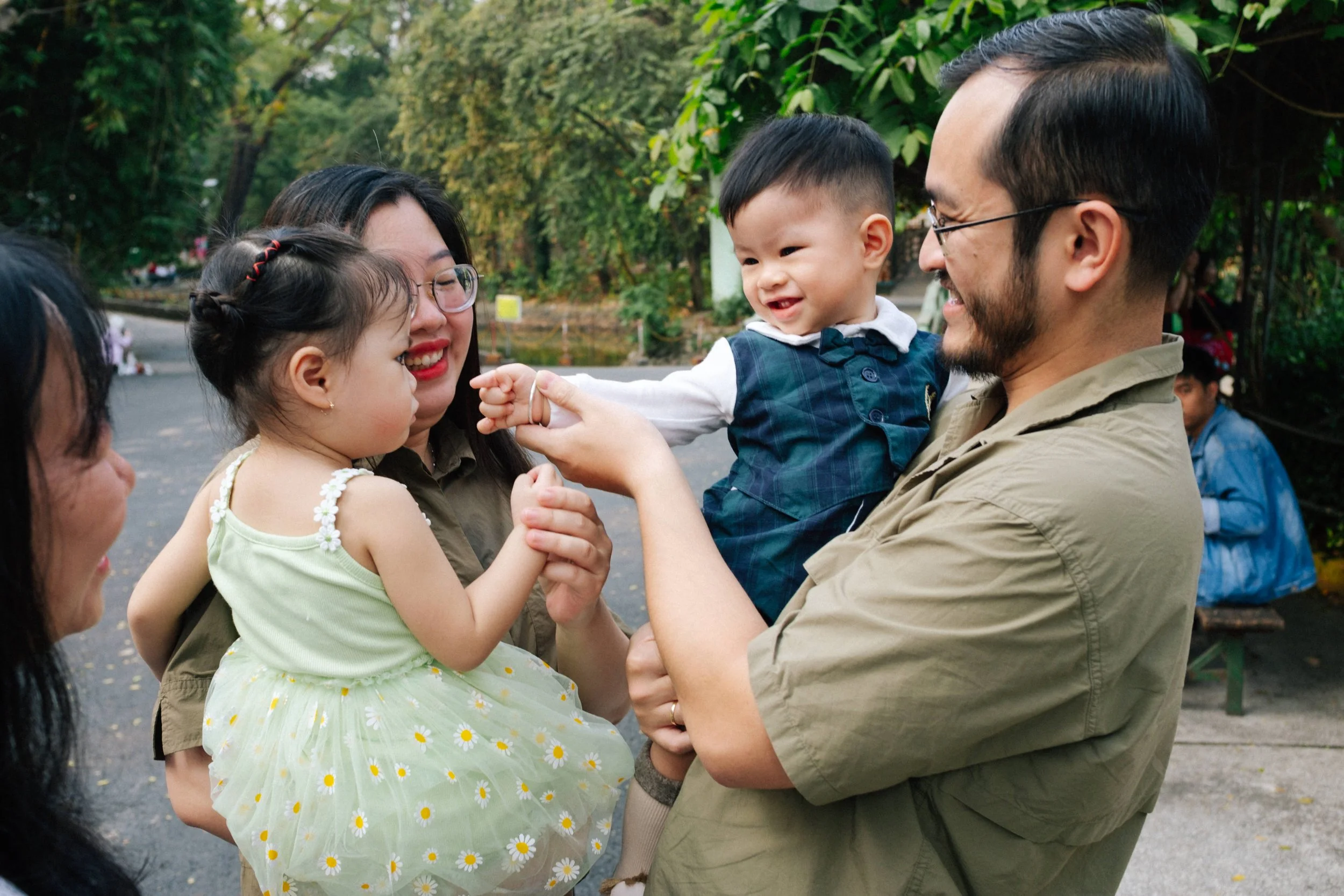 A family of five, including two mothers, a father, a little girl, and a little boy, enjoying a playful moment outdoors in a park with green trees in the background. The father is holding the boy, who is reaching out to the little girl, while the moth