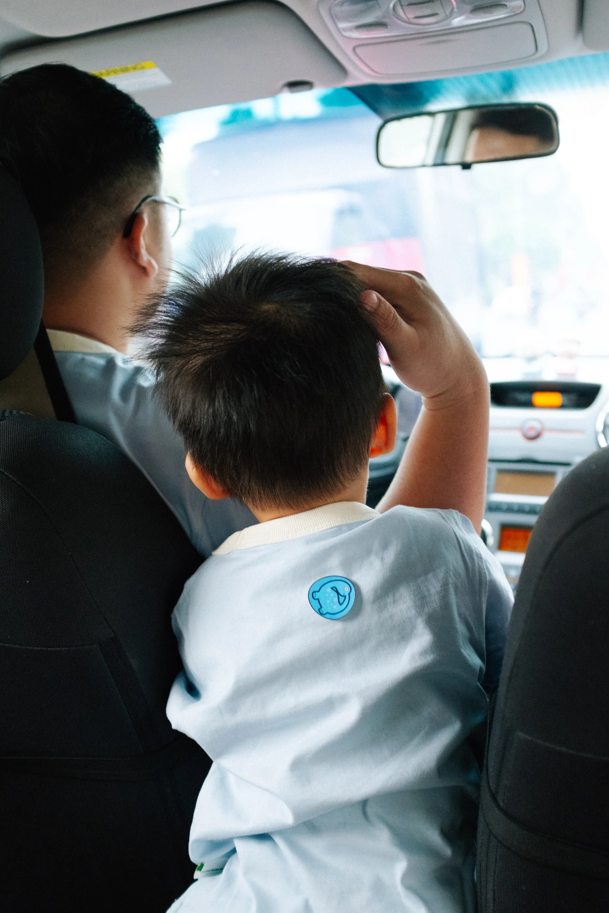 Two children sitting in the backseat of a car, one with glasses and the other scratching their head, both looking forward.