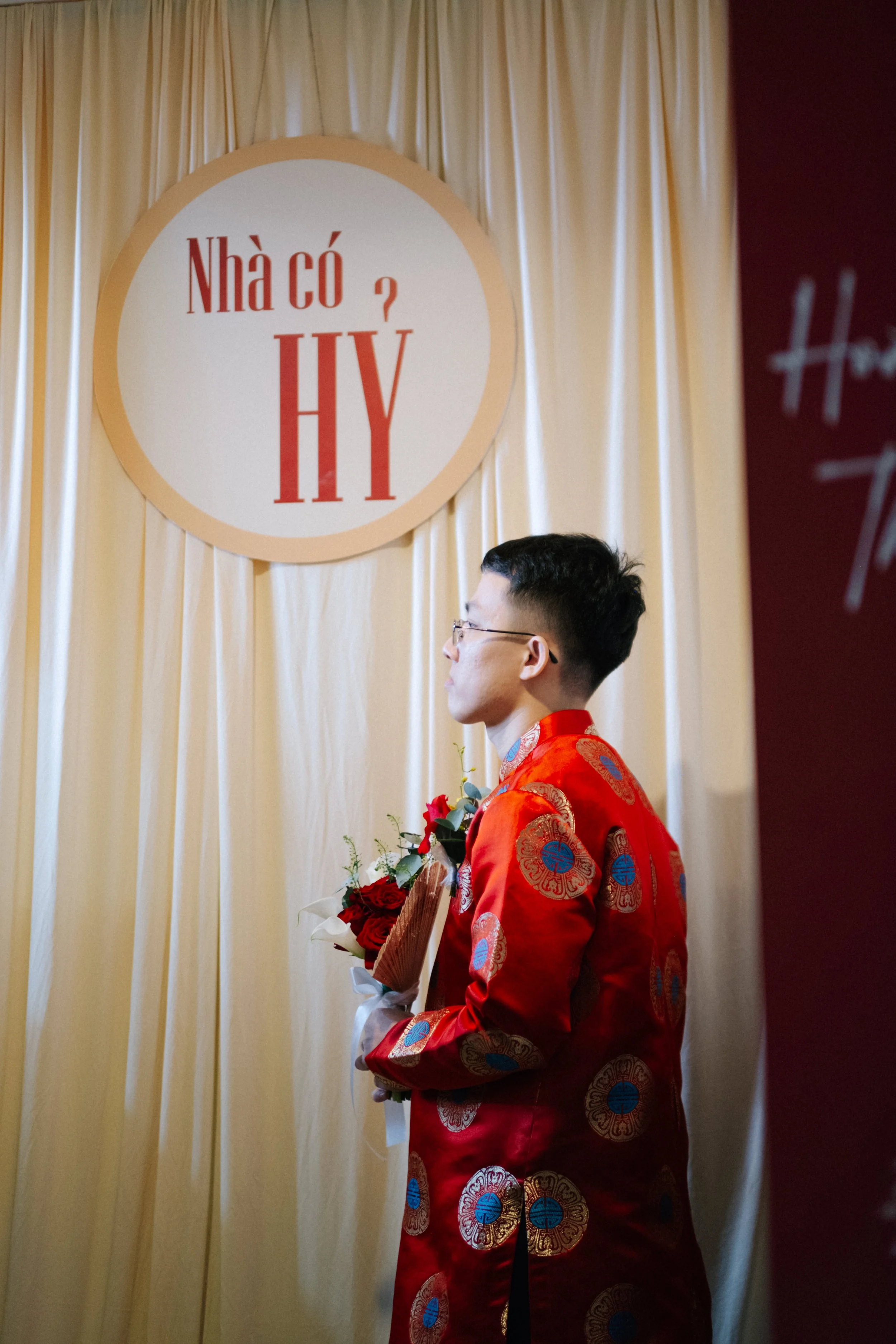 A man dressed in a red traditional Asian garment holding a bouquet of red roses, standing in front of a cream-colored curtain with a large circular sign that has Vietnamese text and the letters 'HY' on it.