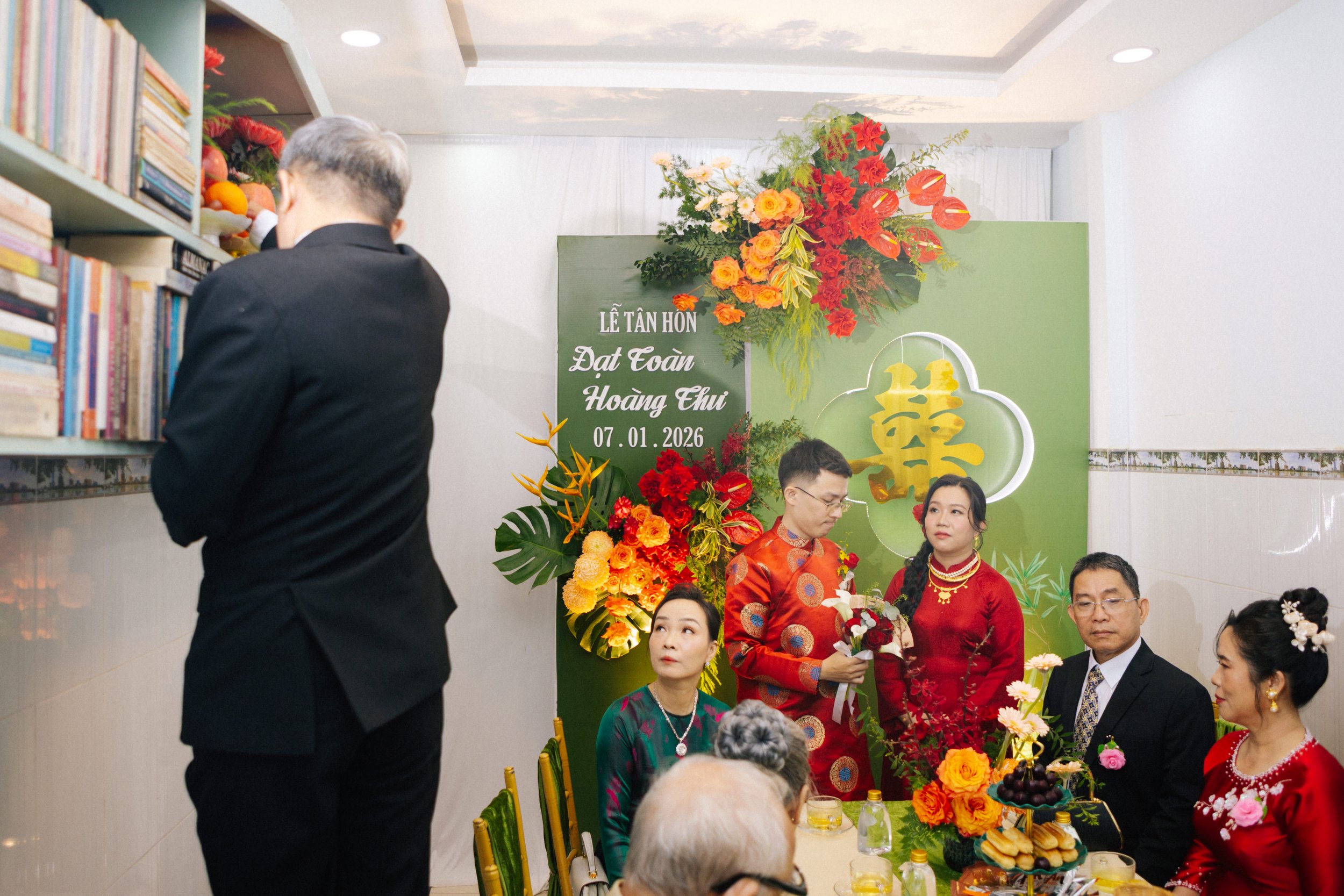 A family gathers for a traditional Vietnamese wedding celebration at a decorated venue with green backdrop and floral arrangements, some individuals are dressed in red traditional attire.