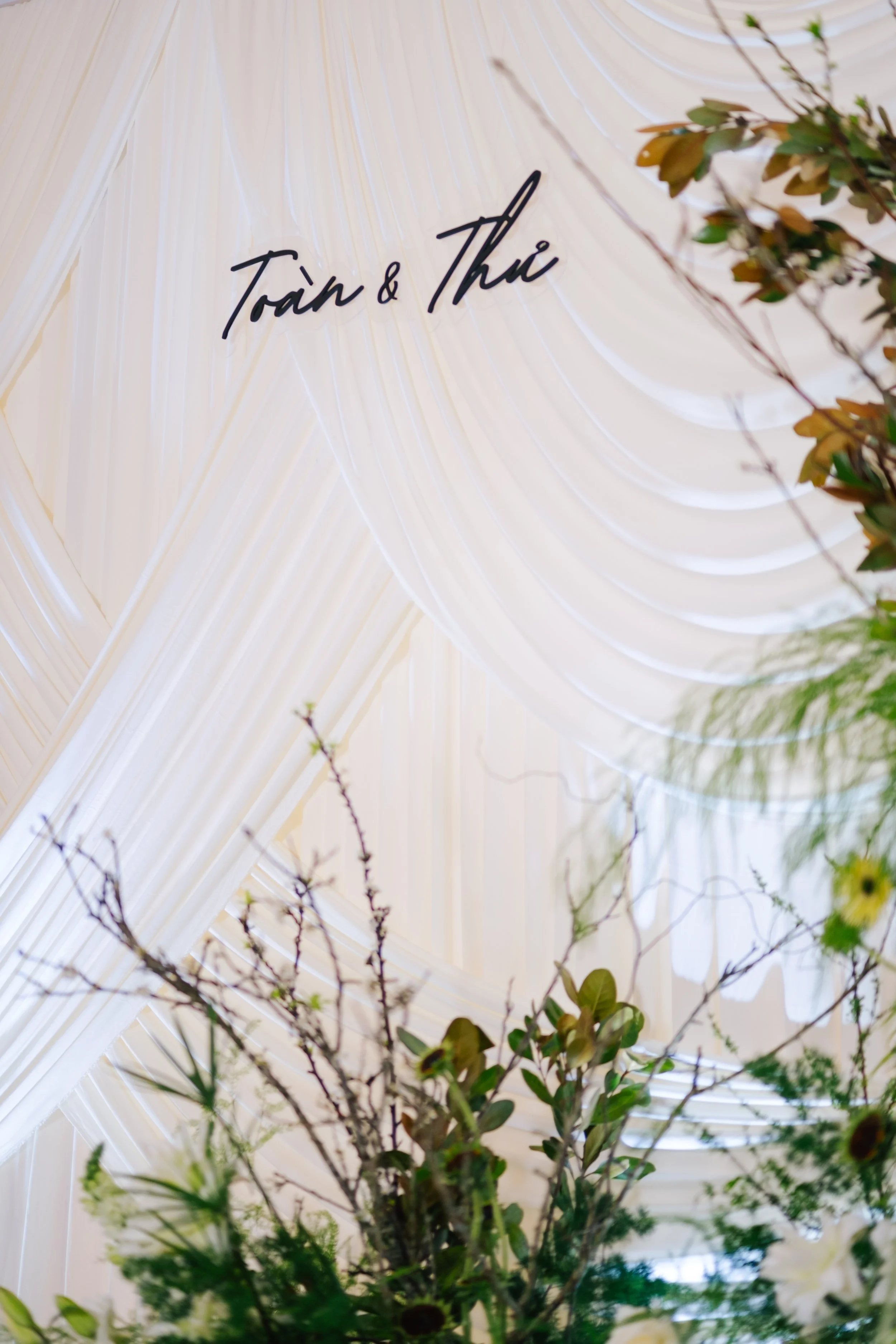Decorative wedding backdrop with white draped fabric and black cursive lettering that reads 'Toàn & Thảo', accented by floral arrangements in the foreground.