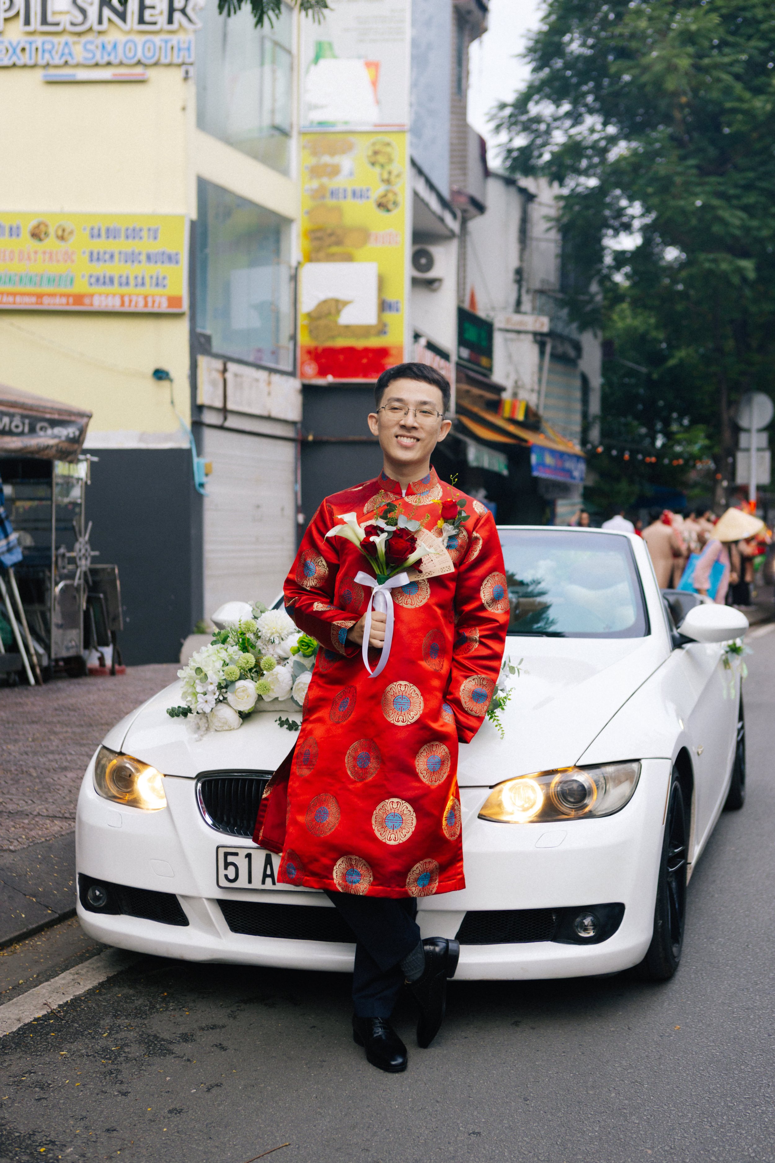 A man in a traditional red Vietnamese wedding áo dài holding a bouquet of red roses, leaning against a white car decorated with flowers on a busy street.