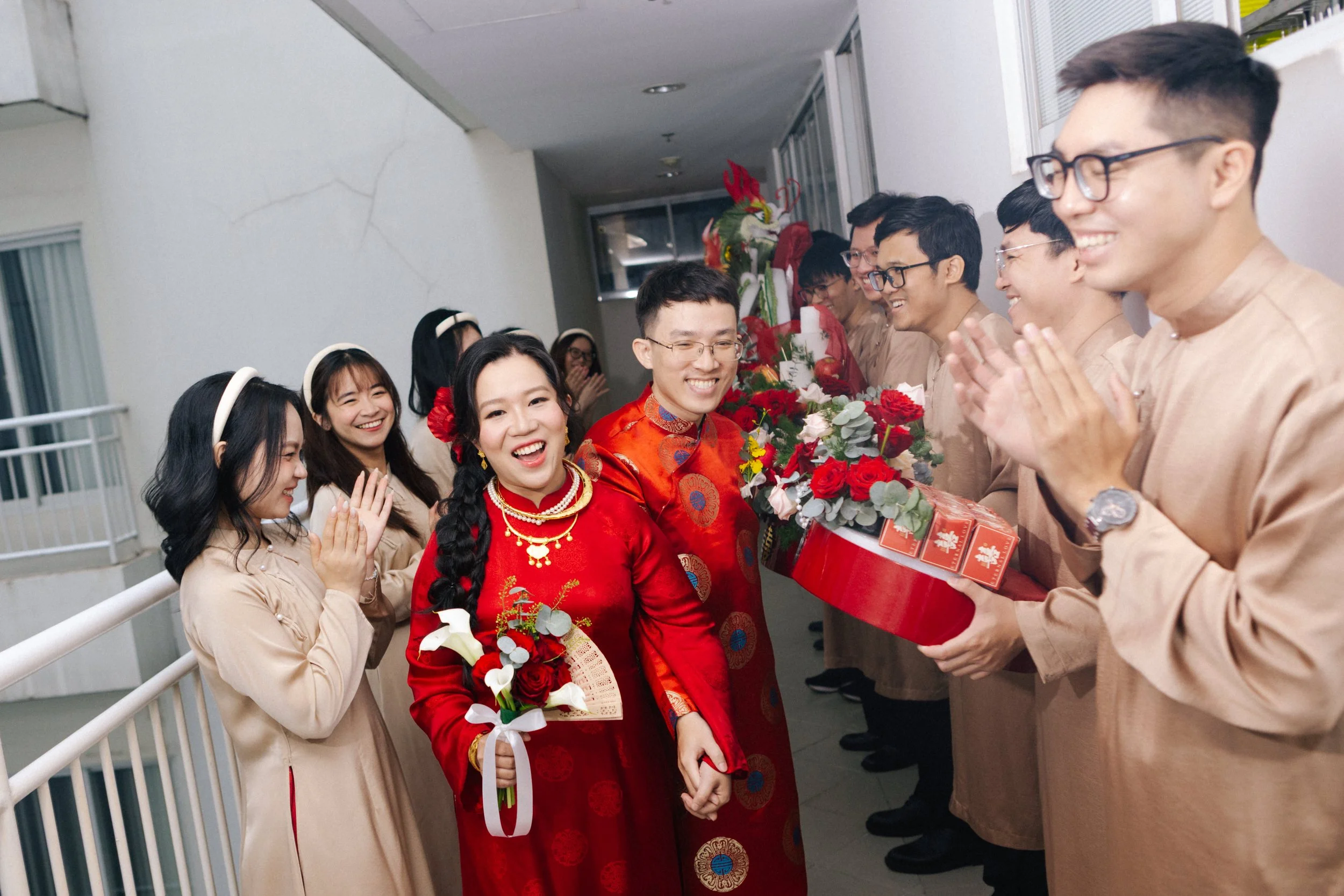 People celebrating at a wedding or cultural event, with women in traditional red dresses and men in beige outfits, surrounded by flower arrangements and gifts, smiling and clapping.