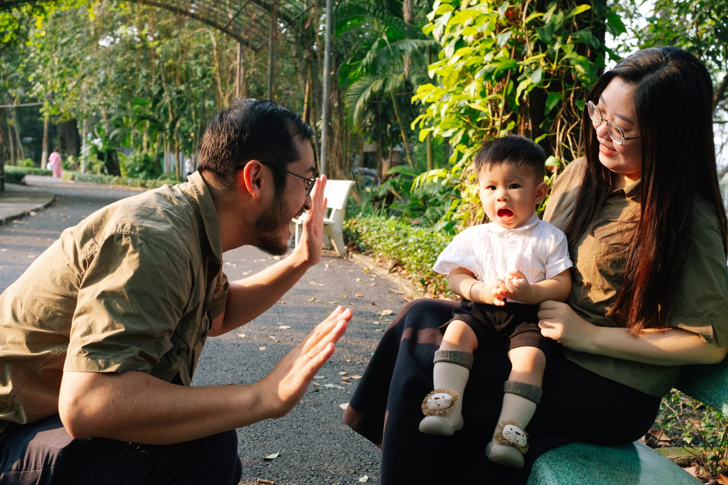 A man playing peek-a-boo with a young child who is sitting on a woman's lap outdoors in a park, surrounded by greenery.