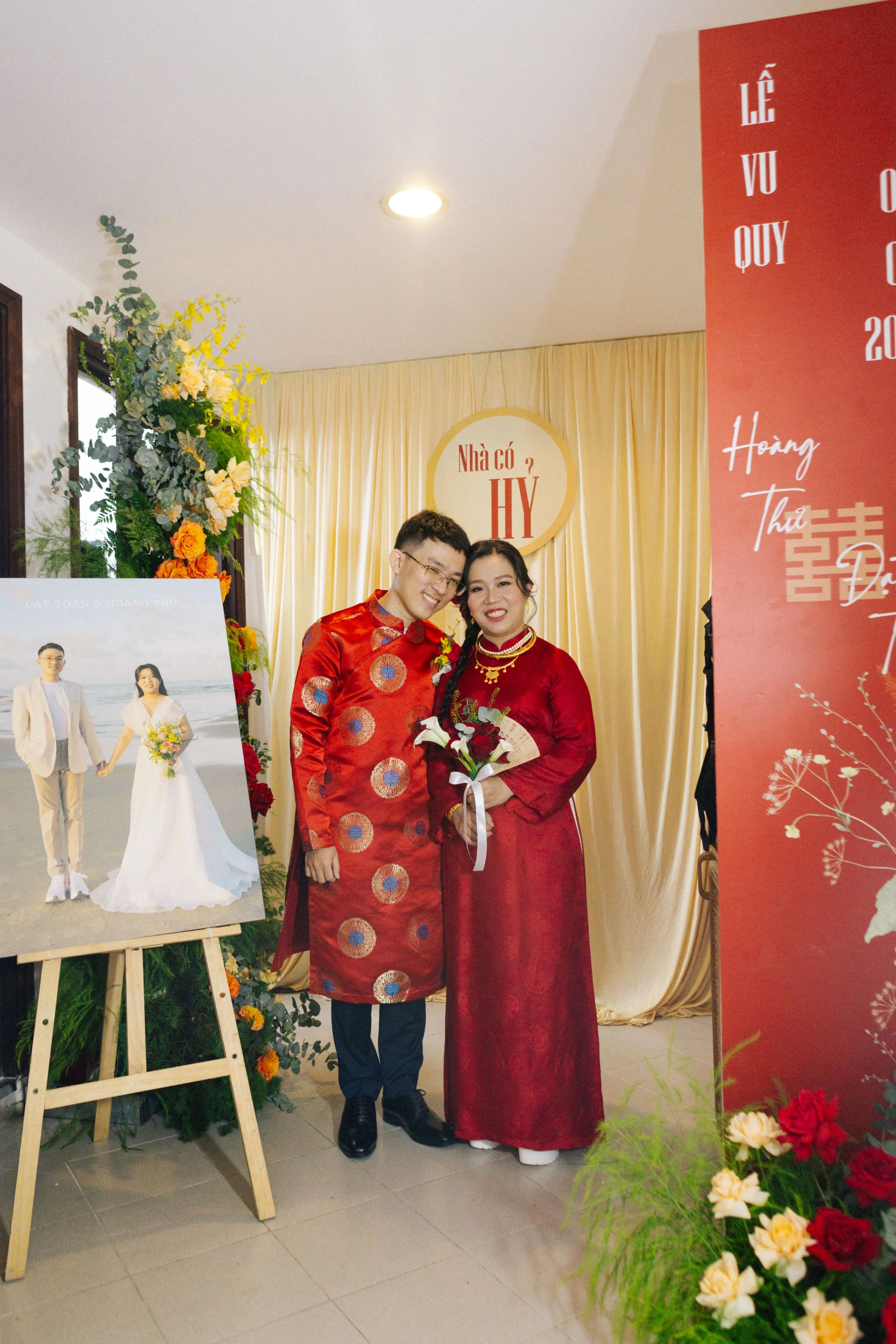 A couple dressed in traditional red Vietnamese attire, standing close together and smiling at a wedding event, with a wedding photograph on an easel and floral decorations around.