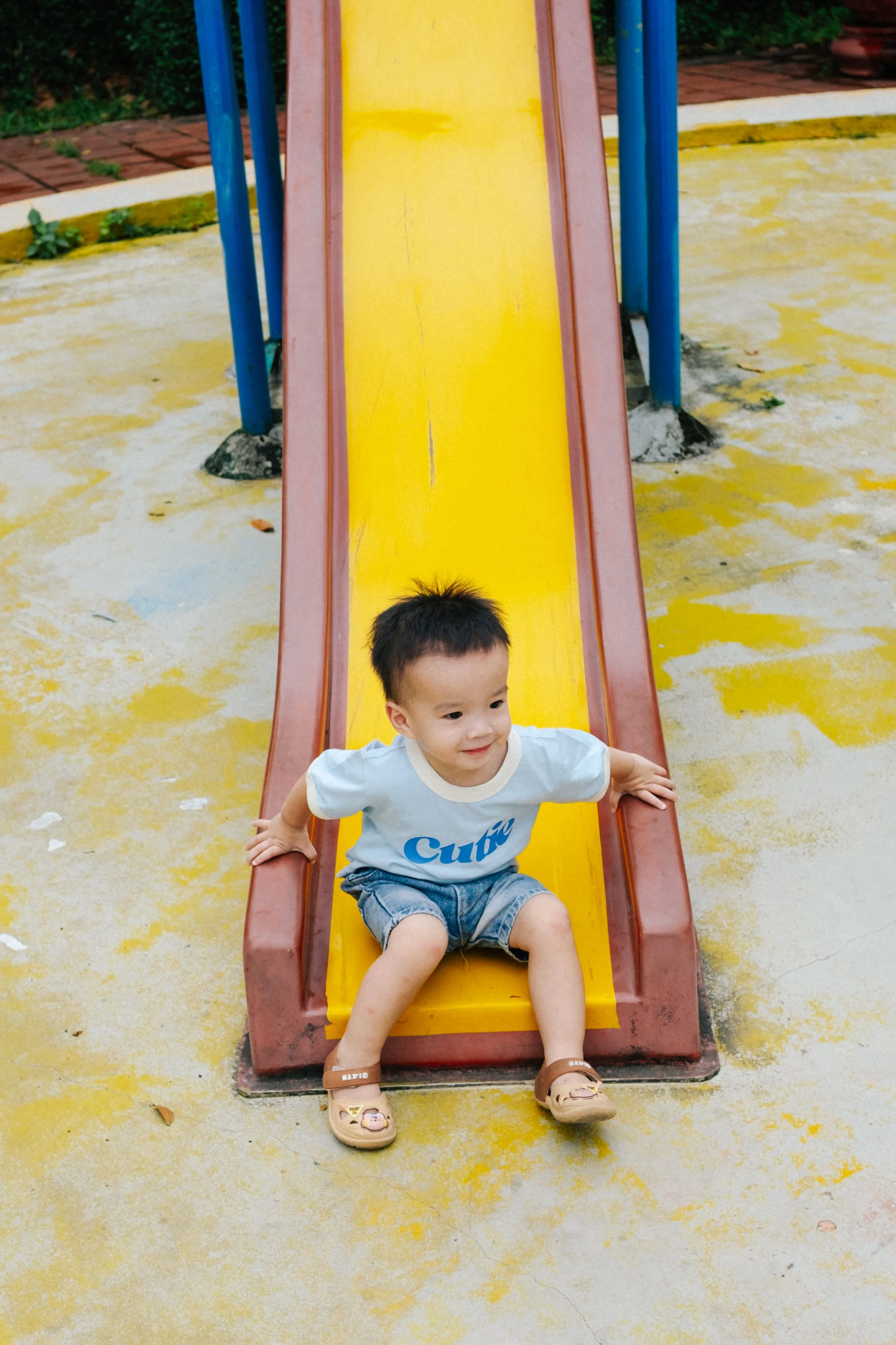Young child sitting at the bottom of a yellow slide at a playground, smiling and looking to the side.