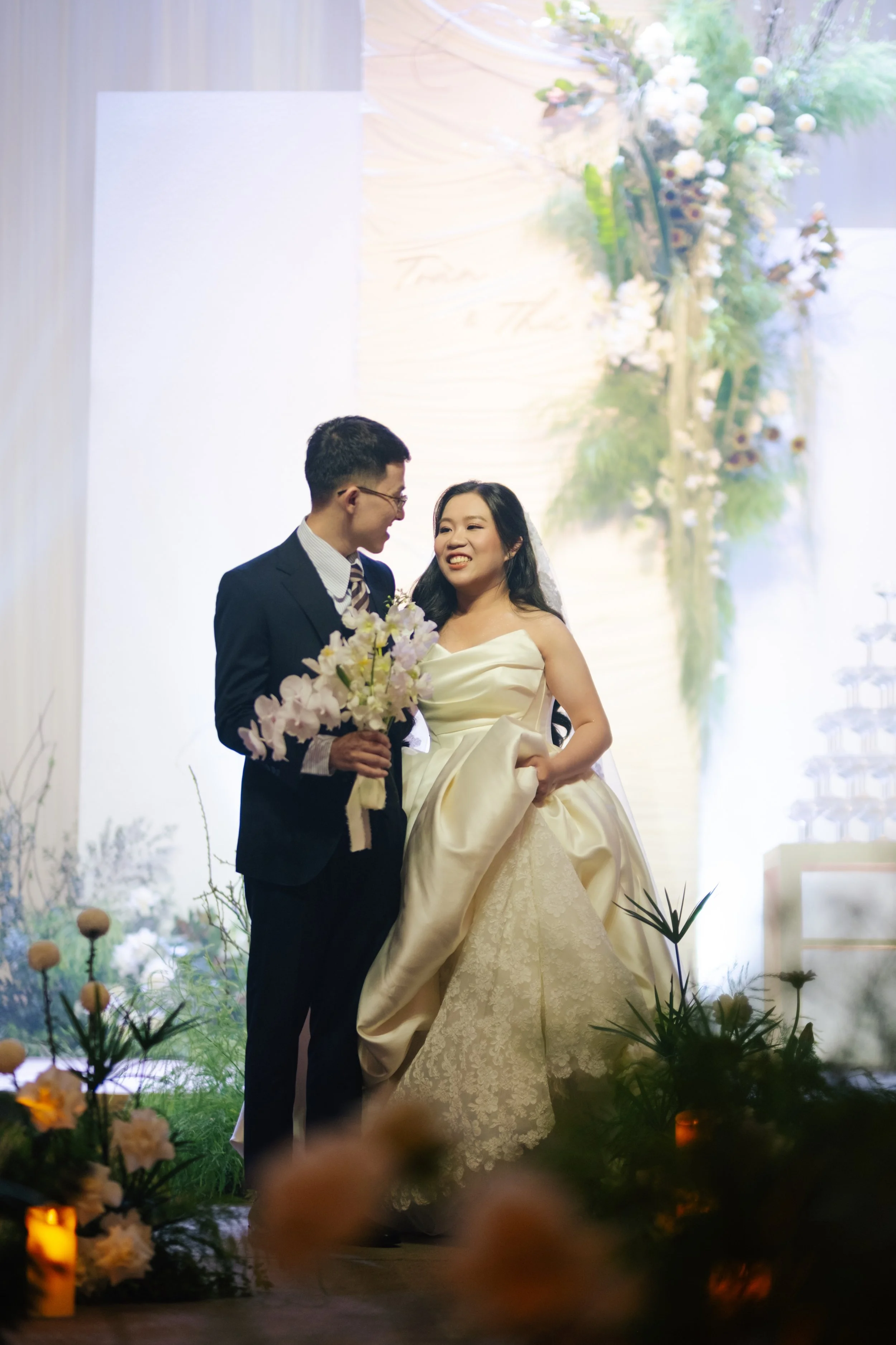 A bride and groom standing on a stage, smiling at each other during their wedding ceremony. The bride is holding her wedding dress with one hand, and the groom is holding a bouquet of flowers. The background features floral decorations and a softly l