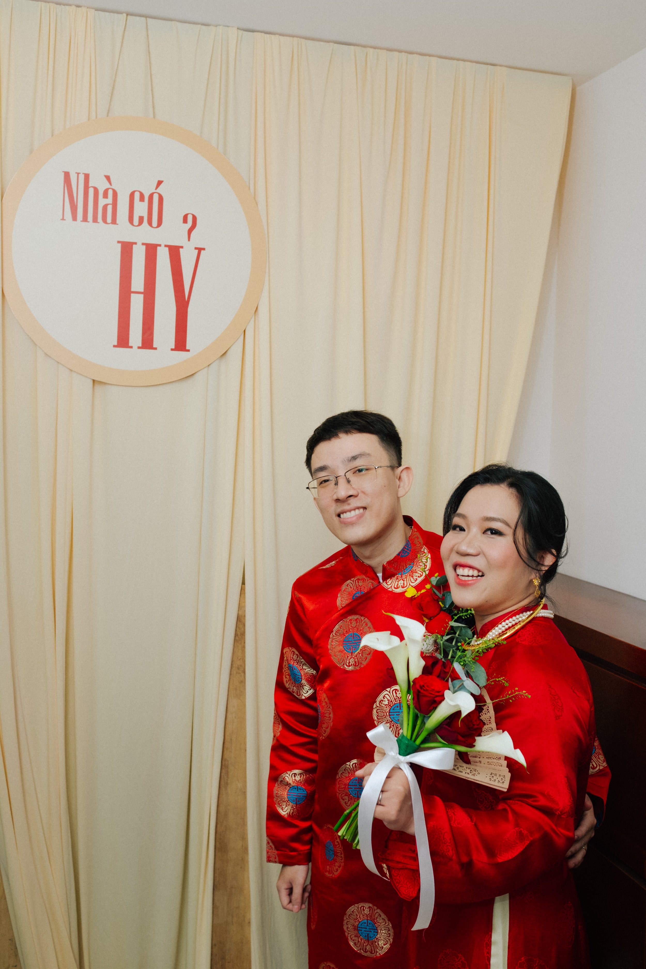 A couple in red traditional wedding attire posing indoors, with a woman holding a bouquet of flowers. Behind them, a beige curtain and a sign with Vietnamese text that reads 'Nhà có HY'.