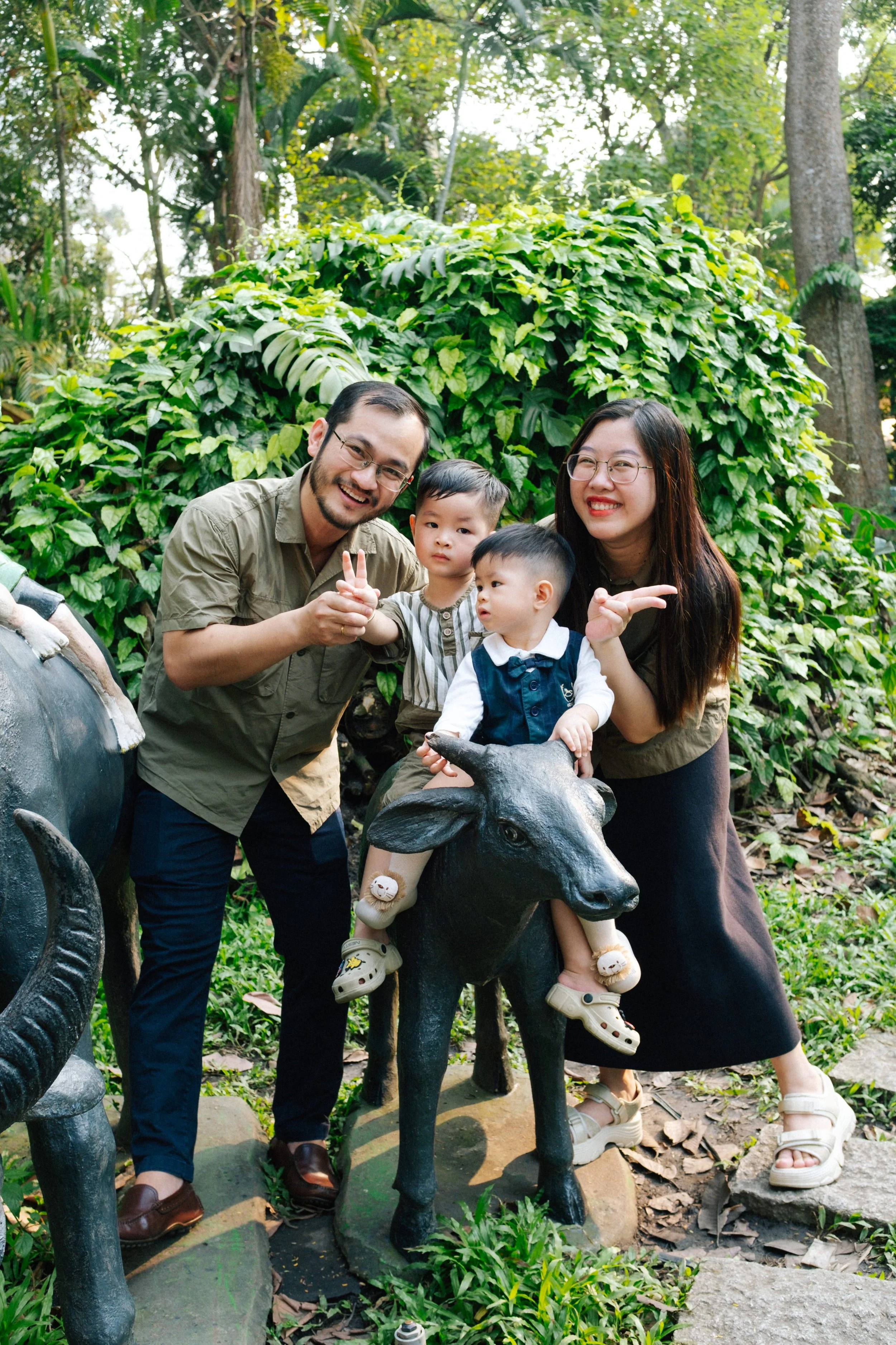 A family of four smiling outdoors in a lush green park, with the children sitting on a black cow statue.