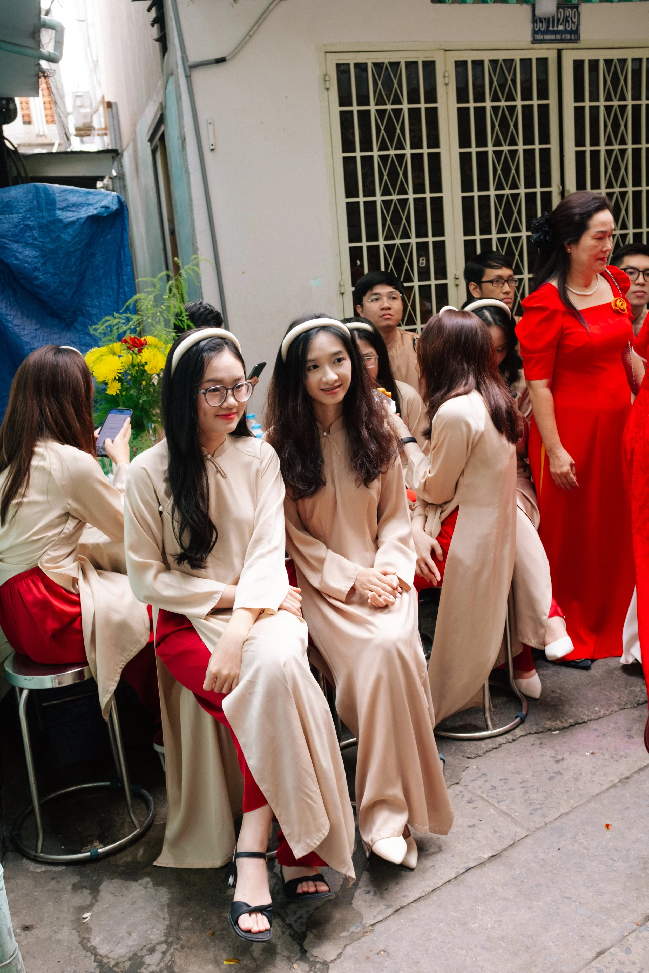 Group of women dressed in traditional outfits, sitting and standing outside near a building with green metal bars on the windows.