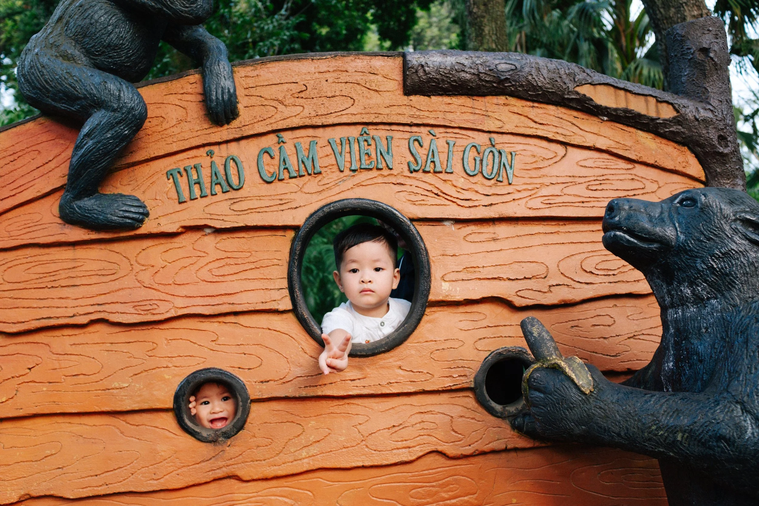 Two children peeking through circular openings in a wooden play structure with animal sculptures, with the sign 'Thảo Cầm Viên Sài Gòn' above
