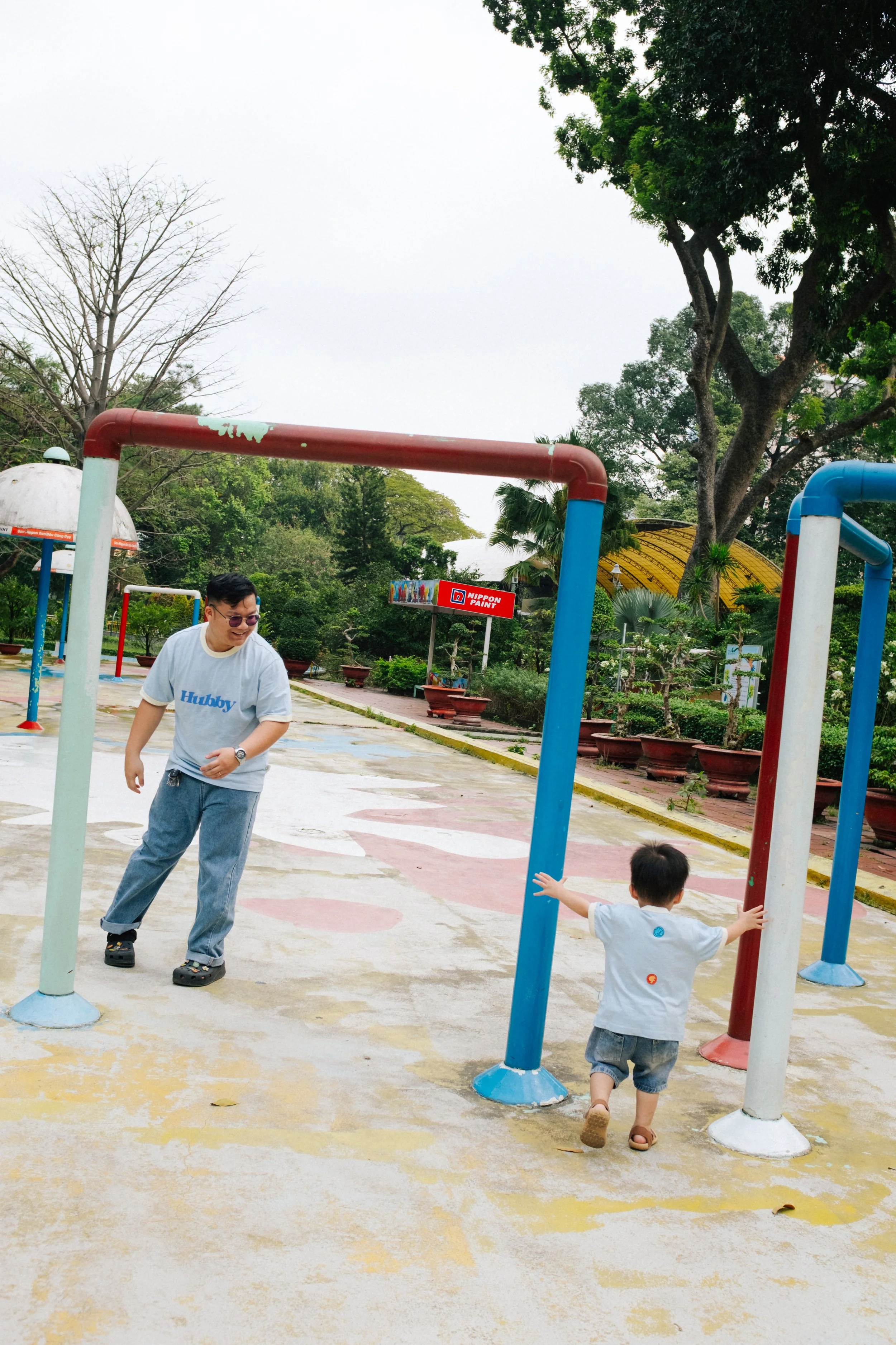 A man and a young boy playing at a park with colorful playground equipment and lush green trees in the background.