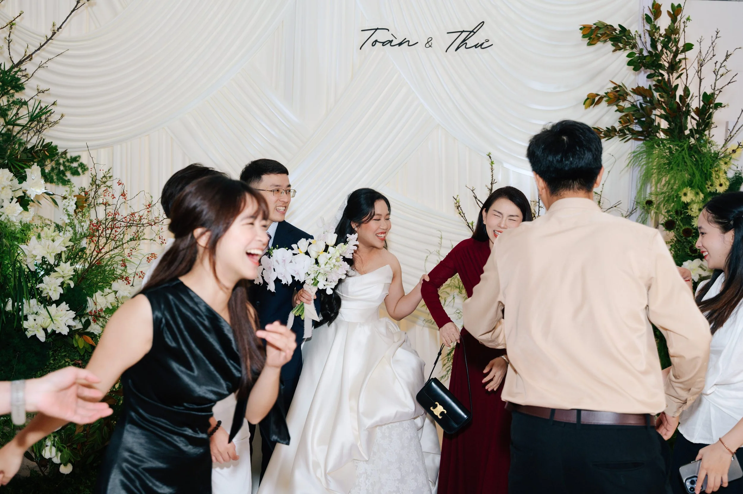 Group of smiling people celebrating a wedding, with the bride in a white dress holding a bouquet, and others dressed in formal attire, standing in front of a decorated backdrop with greenery and white flowers.