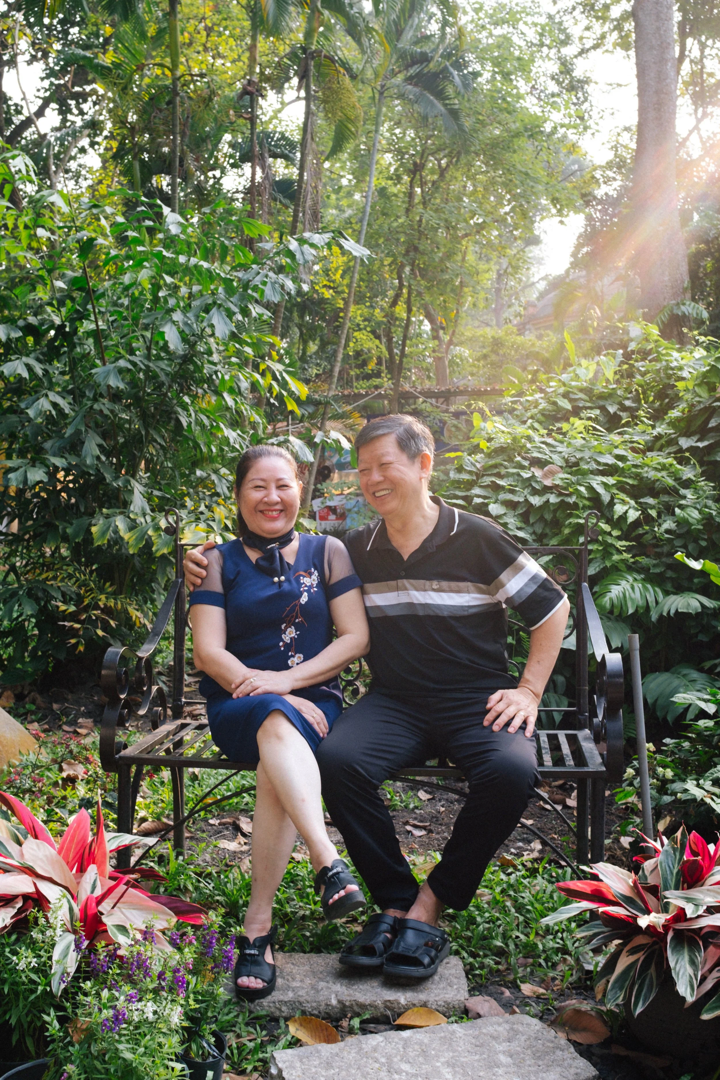 A man and woman sitting on a garden bench, smiling and enjoying each other's company amid lush green foliage and flowers during sunlight.