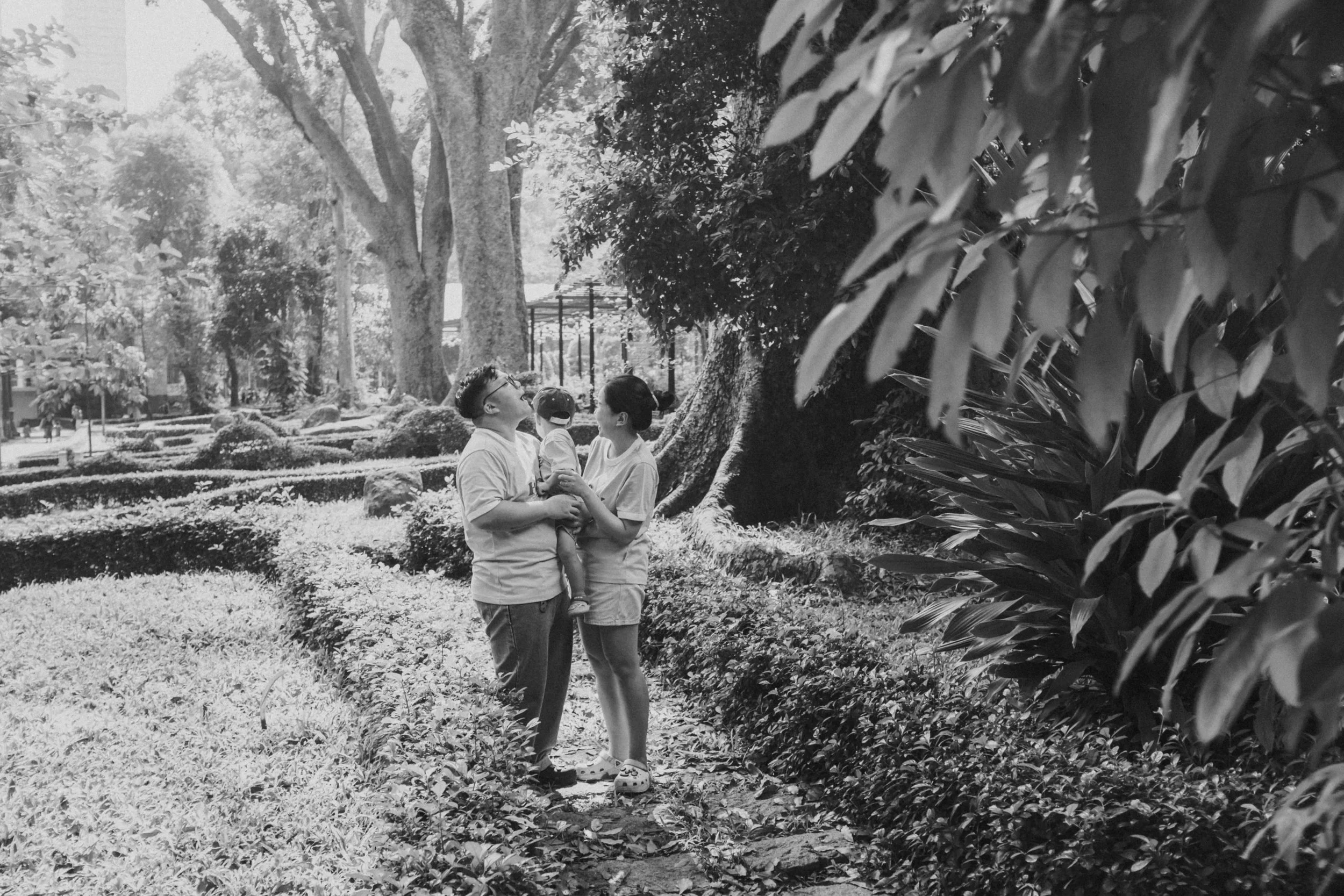 A family of three standing on a garden pathway, with large trees and well-maintained shrubs, sharing a joyful moment outdoors.