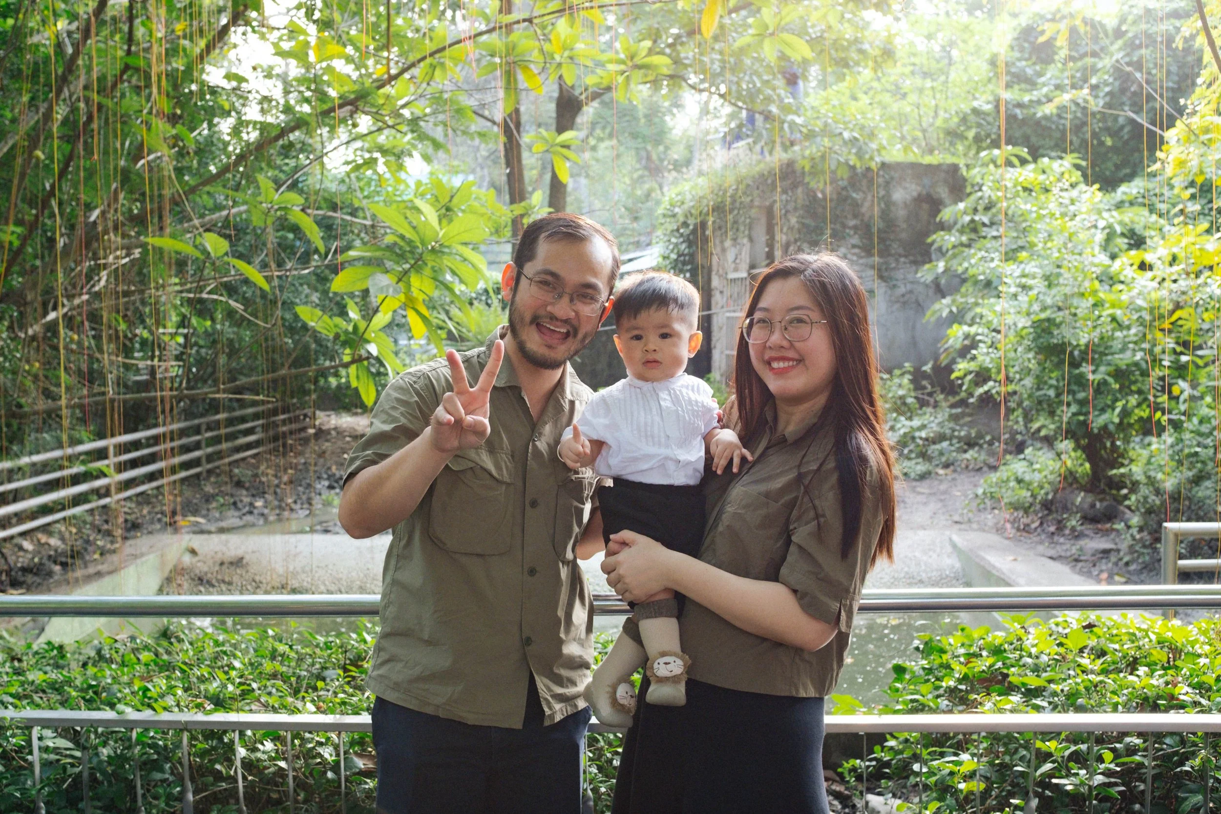 A family of three, a man, woman, and a small girl, posing together outdoors in a lush green environment, with the man displaying a peace sign and everyone smiling.