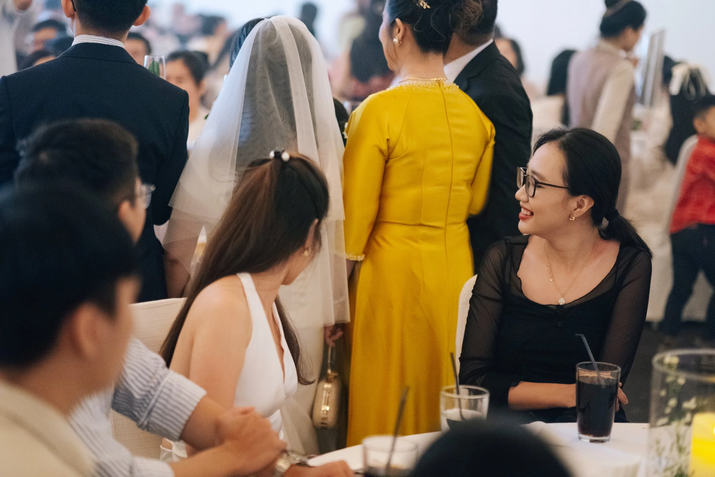 People socializing at a banquet, some seated at a table with drinks, others standing and talking, in an elegant indoor setting.