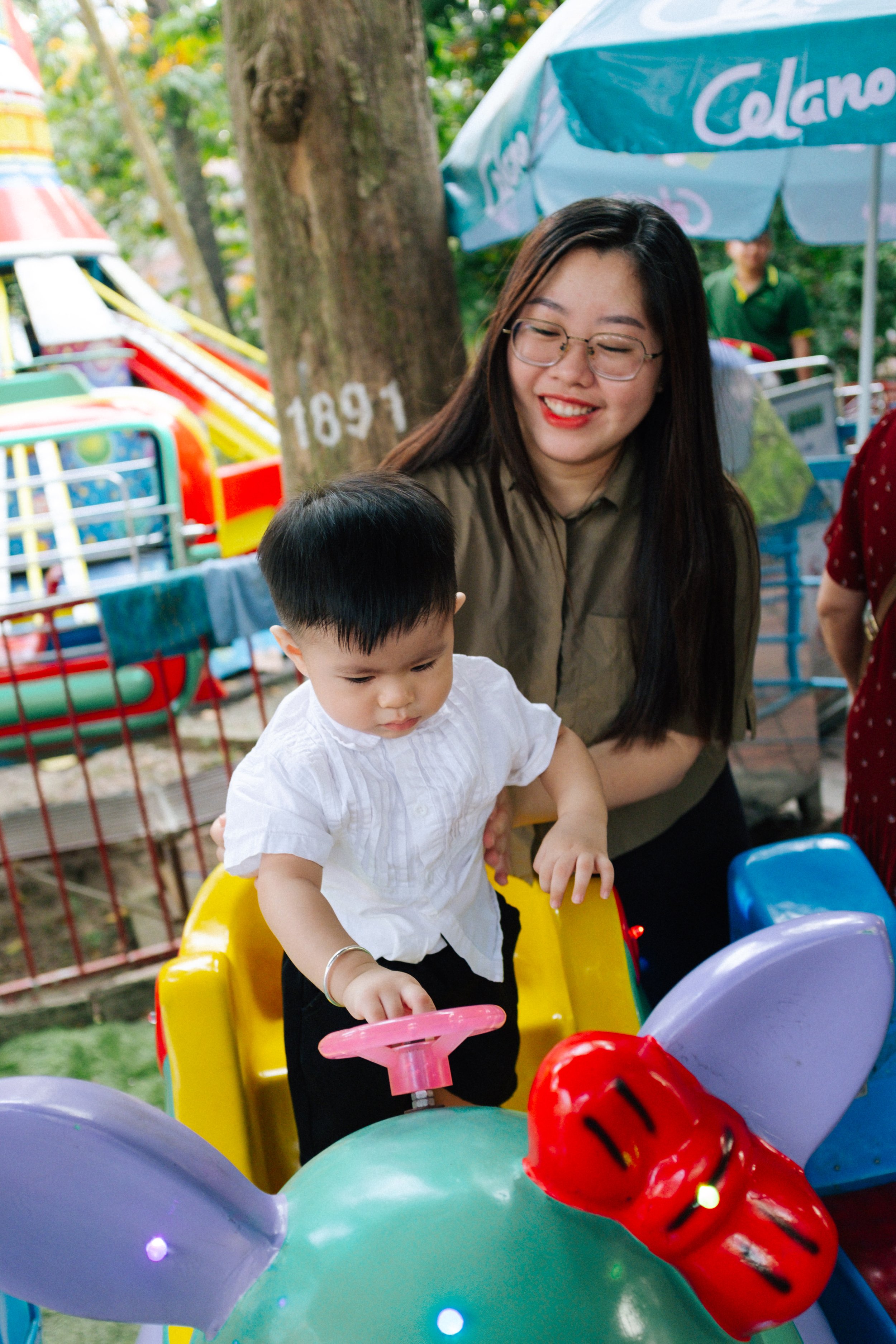 A young child riding a colorful animal-themed ride at an amusement park, guided by a smiling woman with glasses and long dark hair.