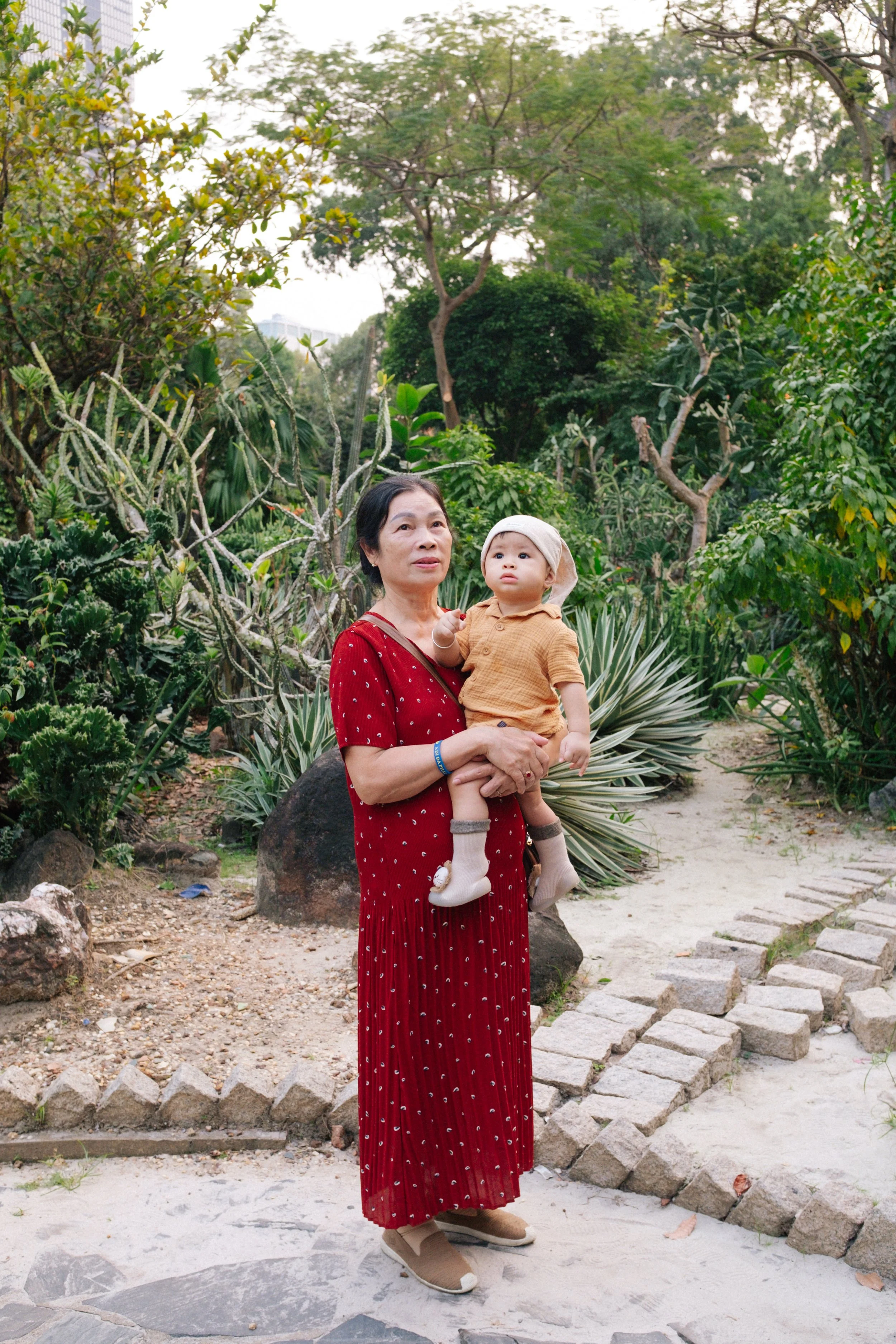 An elderly woman in a red dress holding a young child in an orange outfit and hat in a lush garden with various green plants and trees.