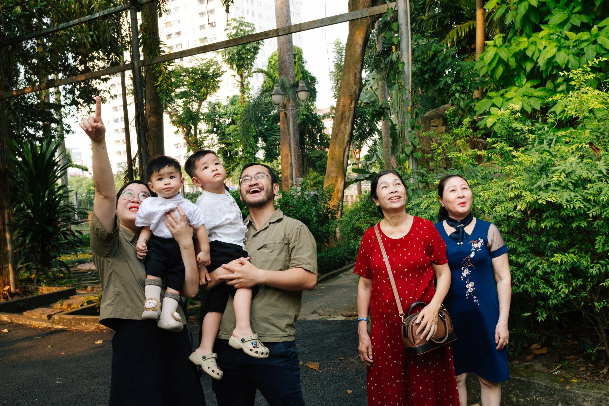 Family photo in a lush park with three generations: woman in olive green shirt and glasses pointing upwards, holding two young boys; man in tan shirt smiling; two women in red and blue dresses observing.