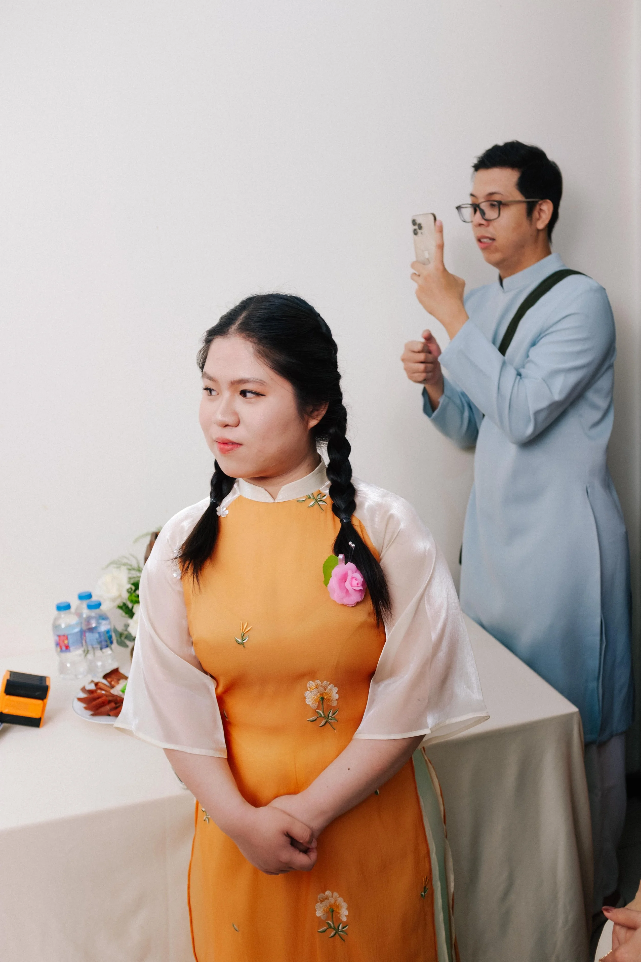 A young woman in a traditional Vietnamese dress with braids stands with her hands clasped, while a man in glasses and a light blue outfit takes a photo of her with his phone.