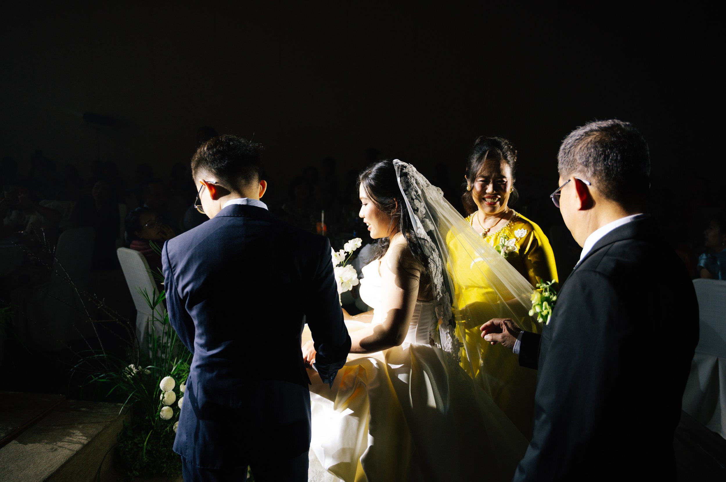 A bride in a white wedding gown and veil, holding a bouquet, is exchanging vows with a groom in a dark suit, while two women in yellow dresses and two men in dark suits look on at a wedding ceremony.