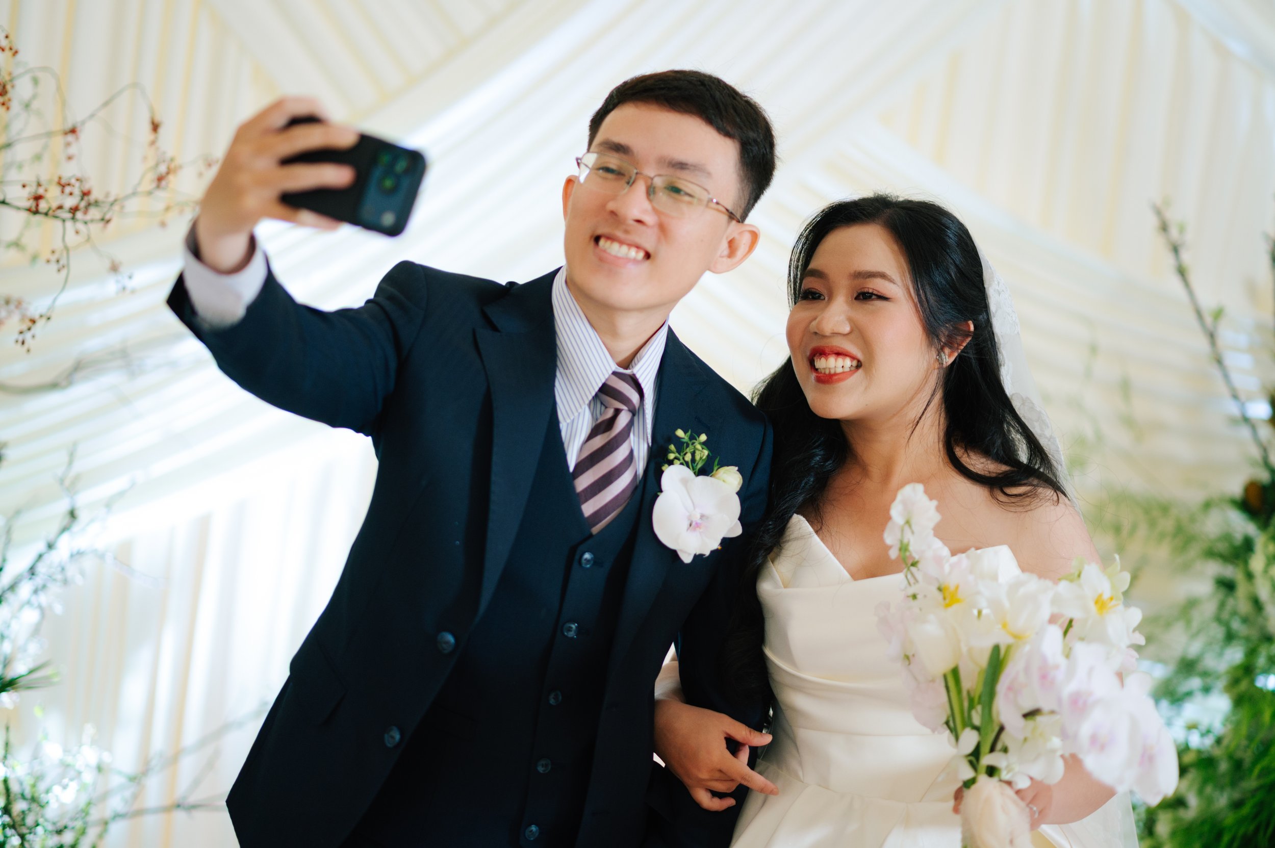 A newlywed couple taking a selfie during their wedding, with the groom in a dark suit and the bride in a white wedding dress holding a bouquet of white flowers.