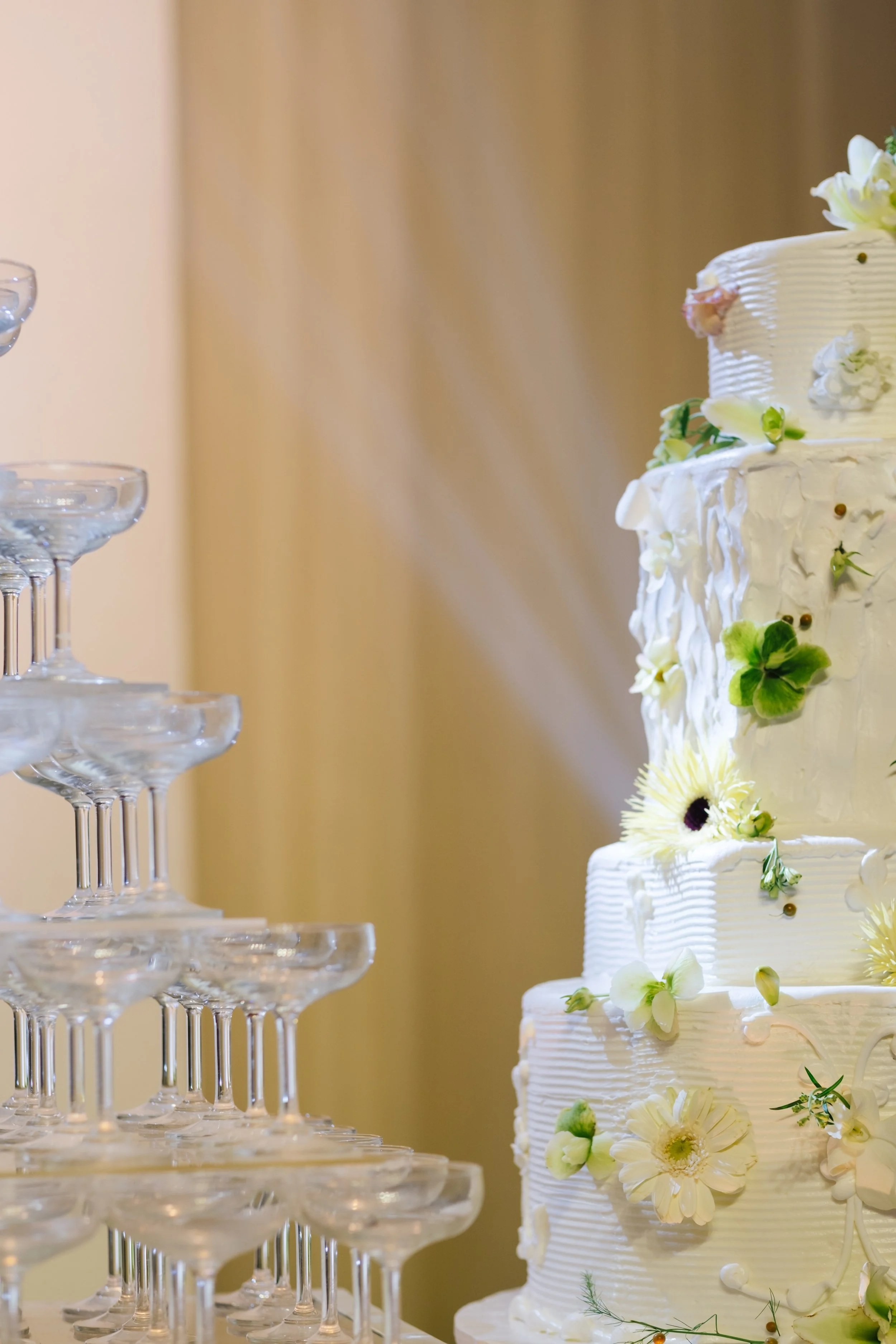 A white wedding cake decorated with flowers next to a pyramid of empty champagne glasses.