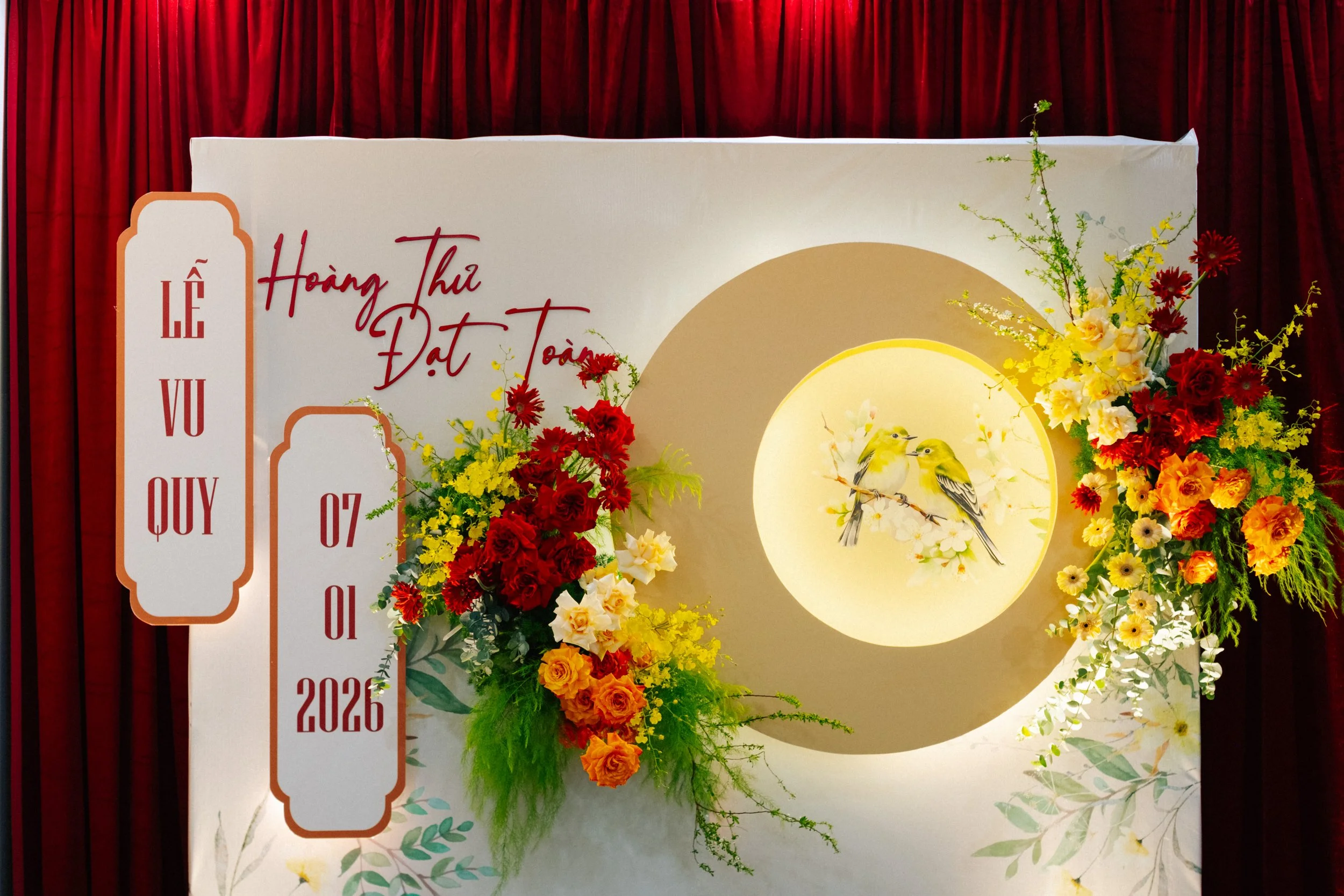 Vietnamese wedding backdrop with floral arrangements, a circular niche with birds, and red curtain background.